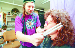 Person with curly hair and glasses receives a hair treatment from a man with dreadlocks in a room.