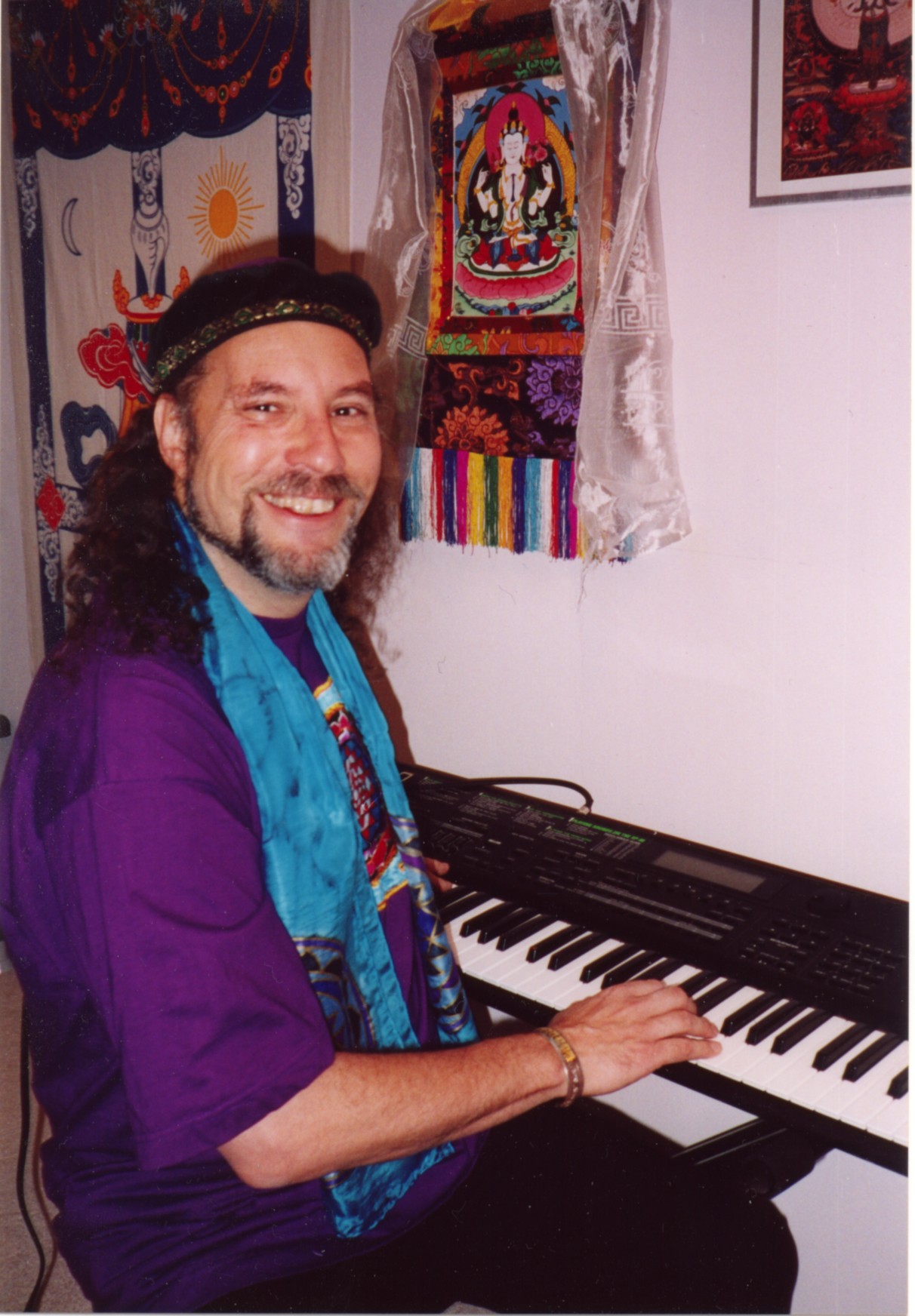 Man with curly hair and beard wearing purple shirt and blue scarf, sitting at keyboard, smiling, colorful textiles on wall