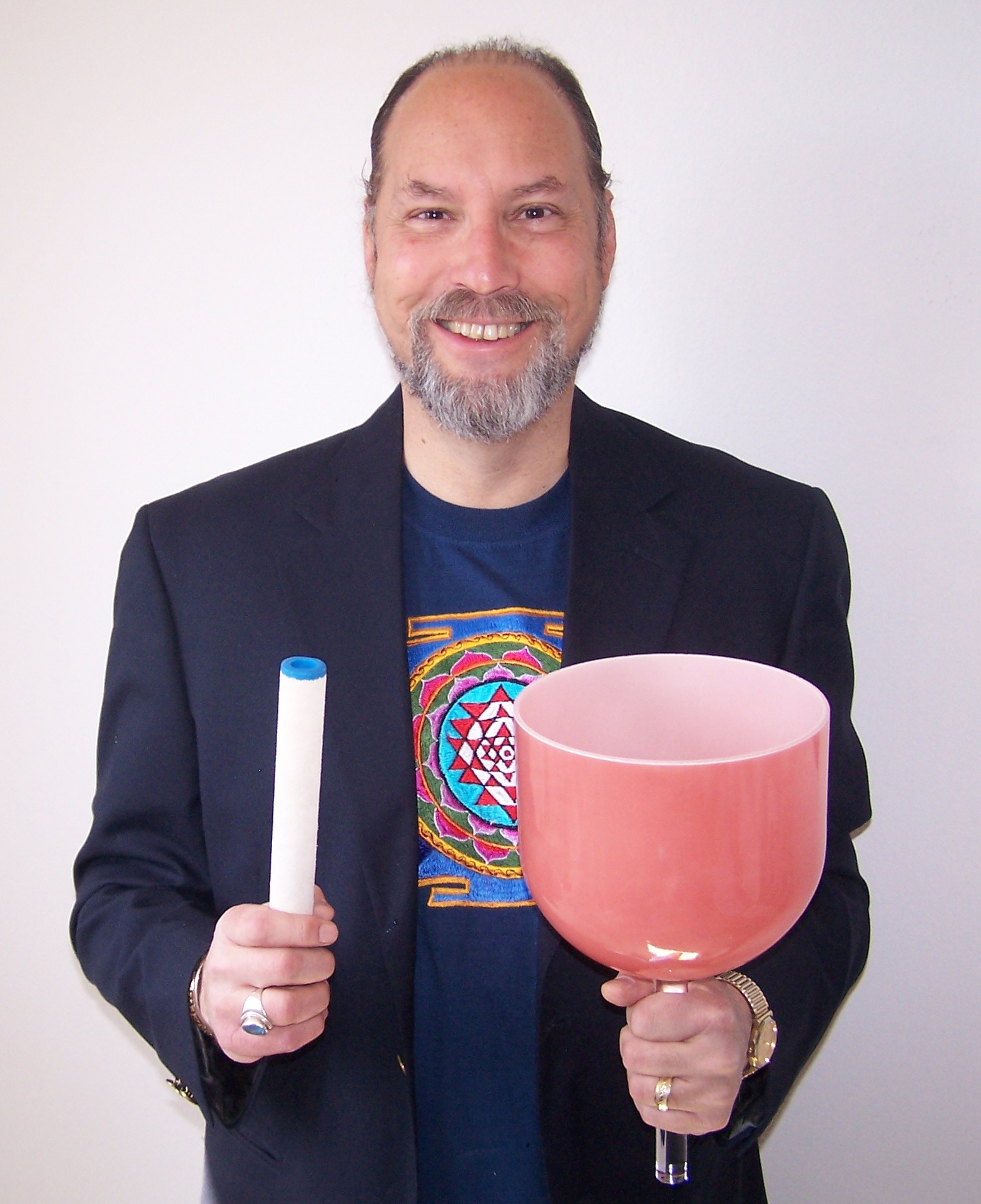 Man with a beard and long hair, smiling, holding a pink singing bowl and a white stick, wearing a dark blazer and colorful shirt.