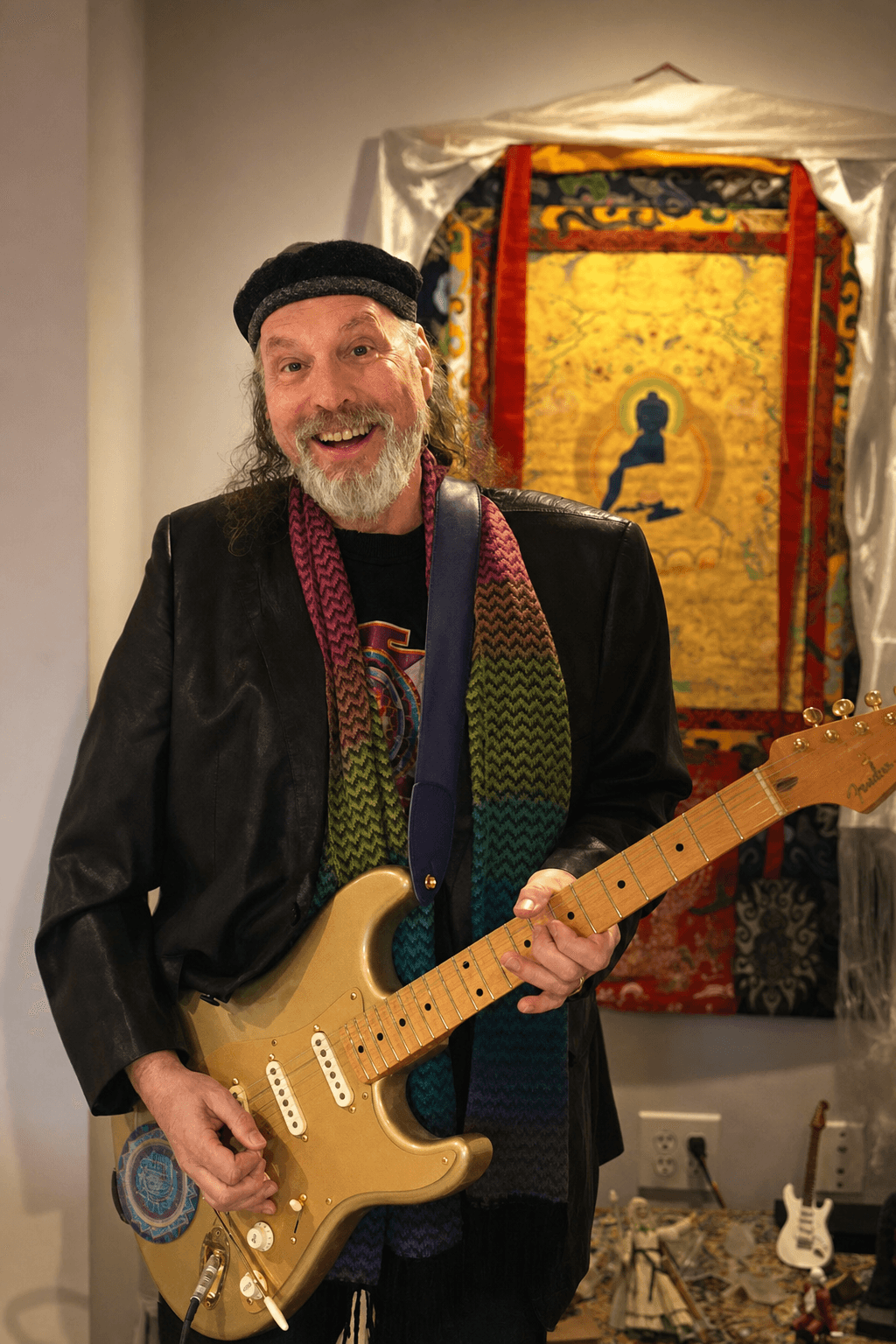 Man with a beard and hat holding an electric guitar, standing in front of a colorful tapestry or artwork.