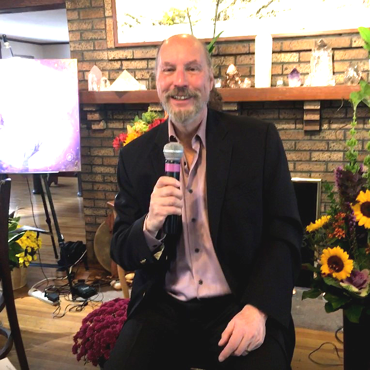 Man with a beard and bald head, wearing a suit, holding a microphone, smiling indoors with flowers and a brick wall.