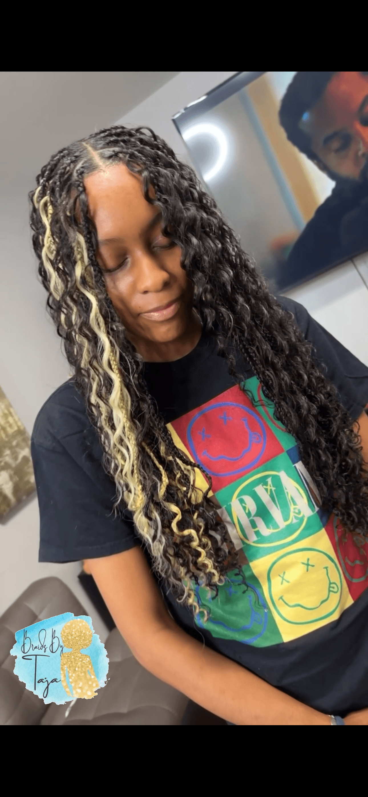 Young girl with long curly hair and blonde highlights, wearing a colorful graphic T-shirt, standing indoors.