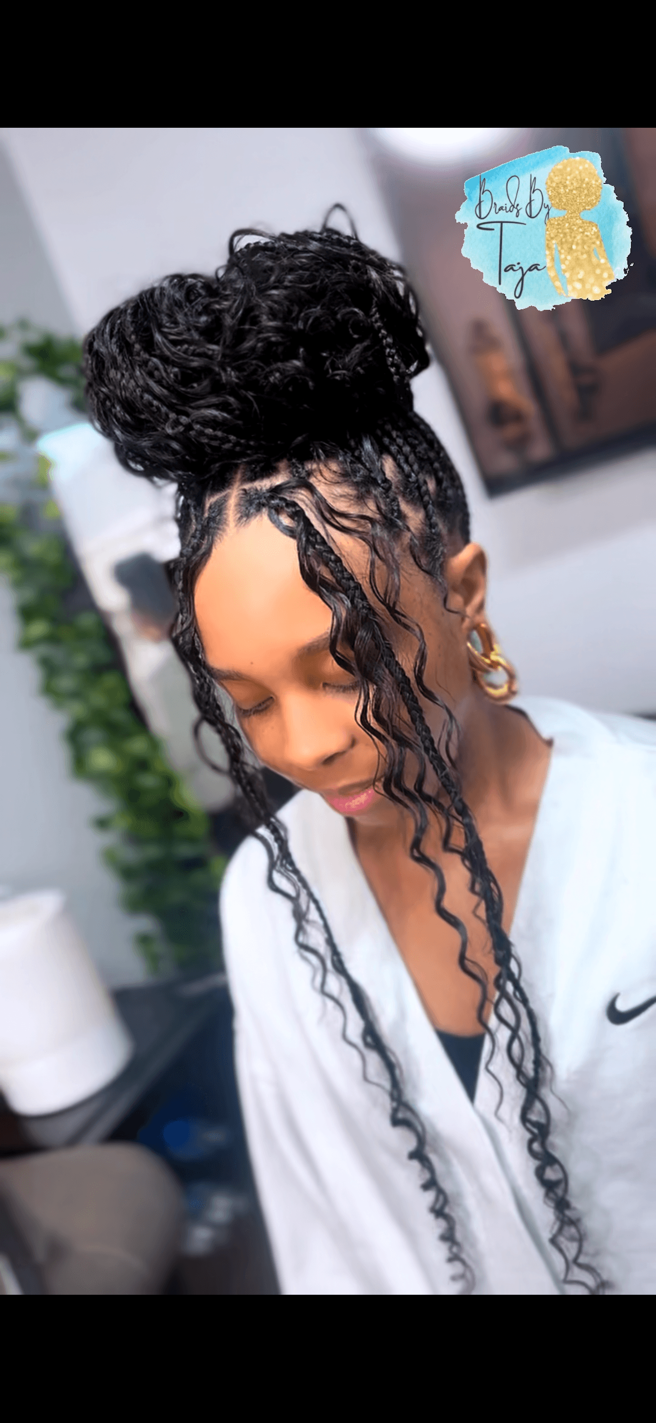 Woman with styled curly hair in a bun, wearing a white Nike top, looking down, indoors with plants and artwork.