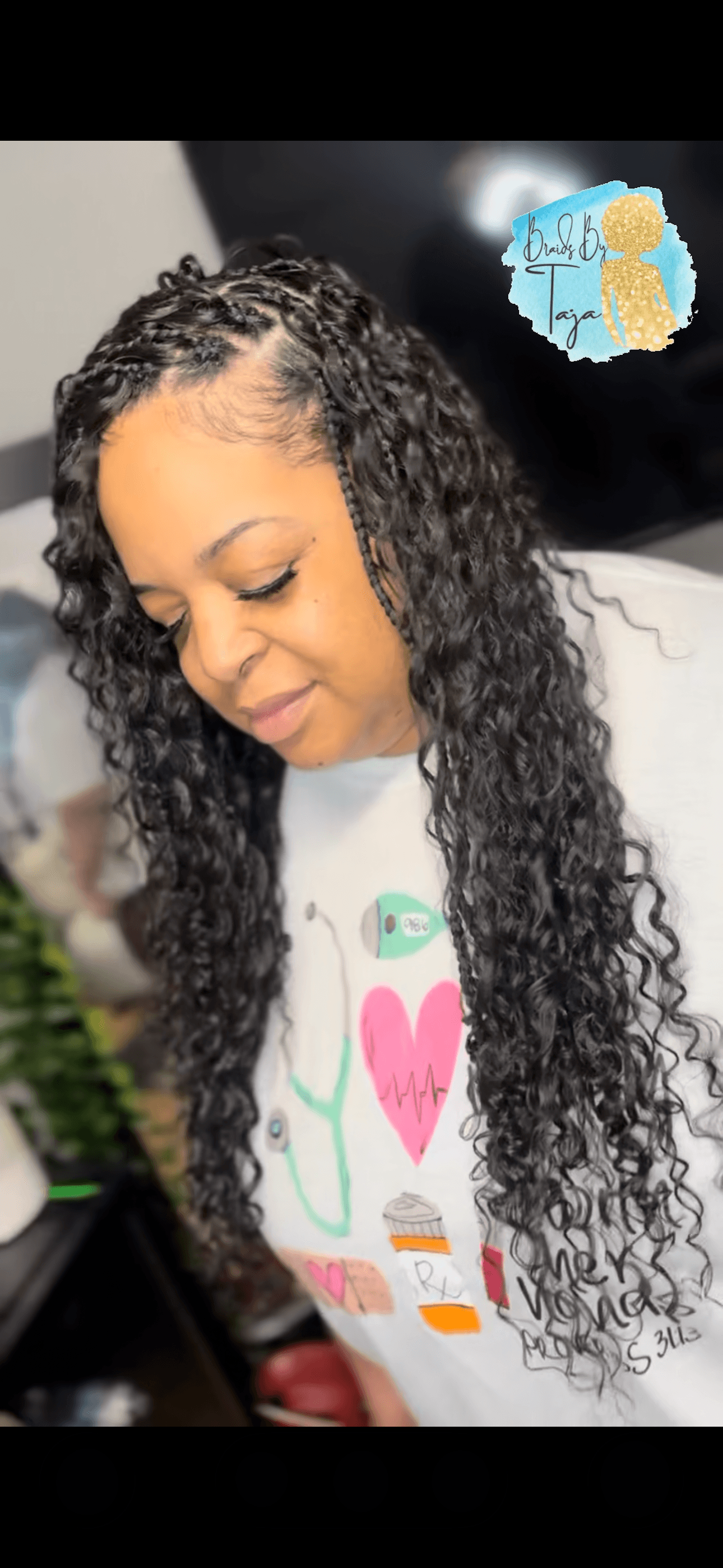Young woman with long, curly hair looking down, wearing a white shirt with colorful designs, in an indoor setting.