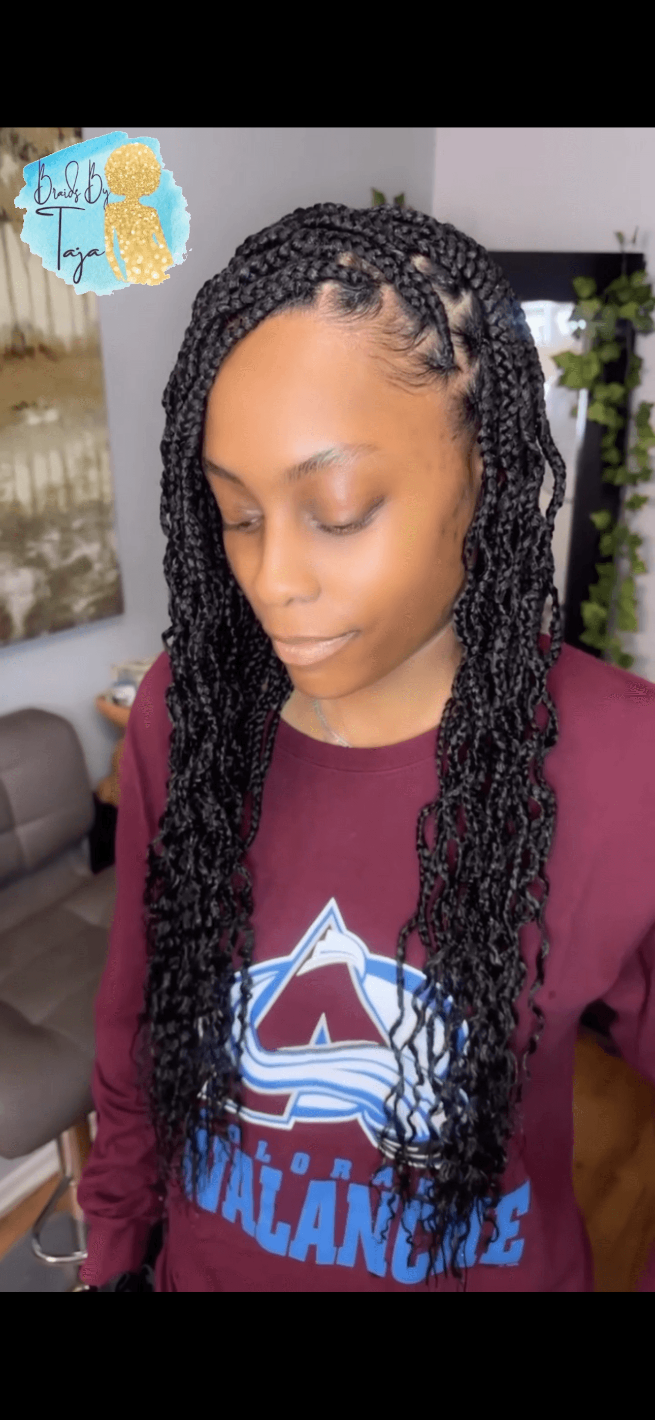 Young woman with braided hair wearing a maroon shirt with a graphic design, indoors with plants in the background.