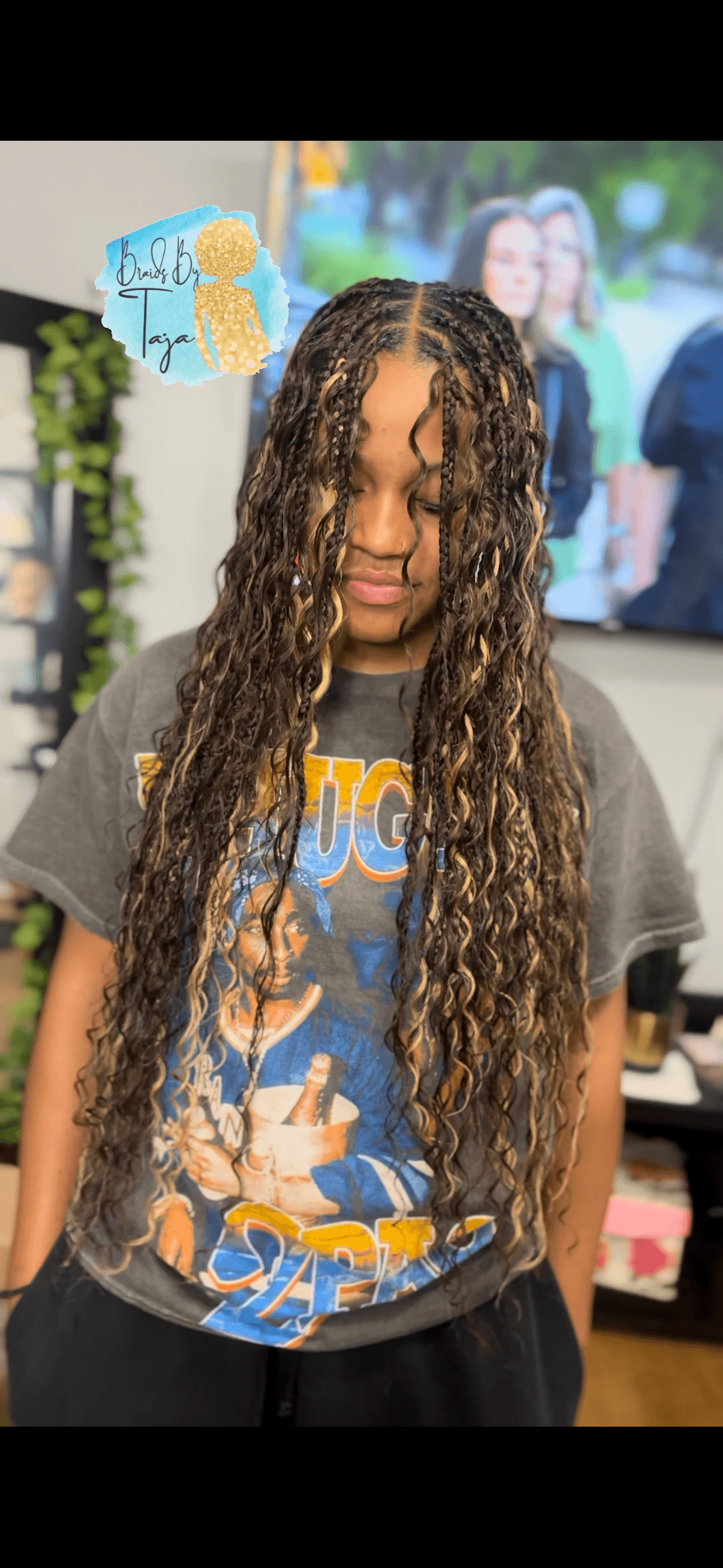 Young person with long curly hair wearing a graphic T-shirt, standing indoors with a TV and people in background.