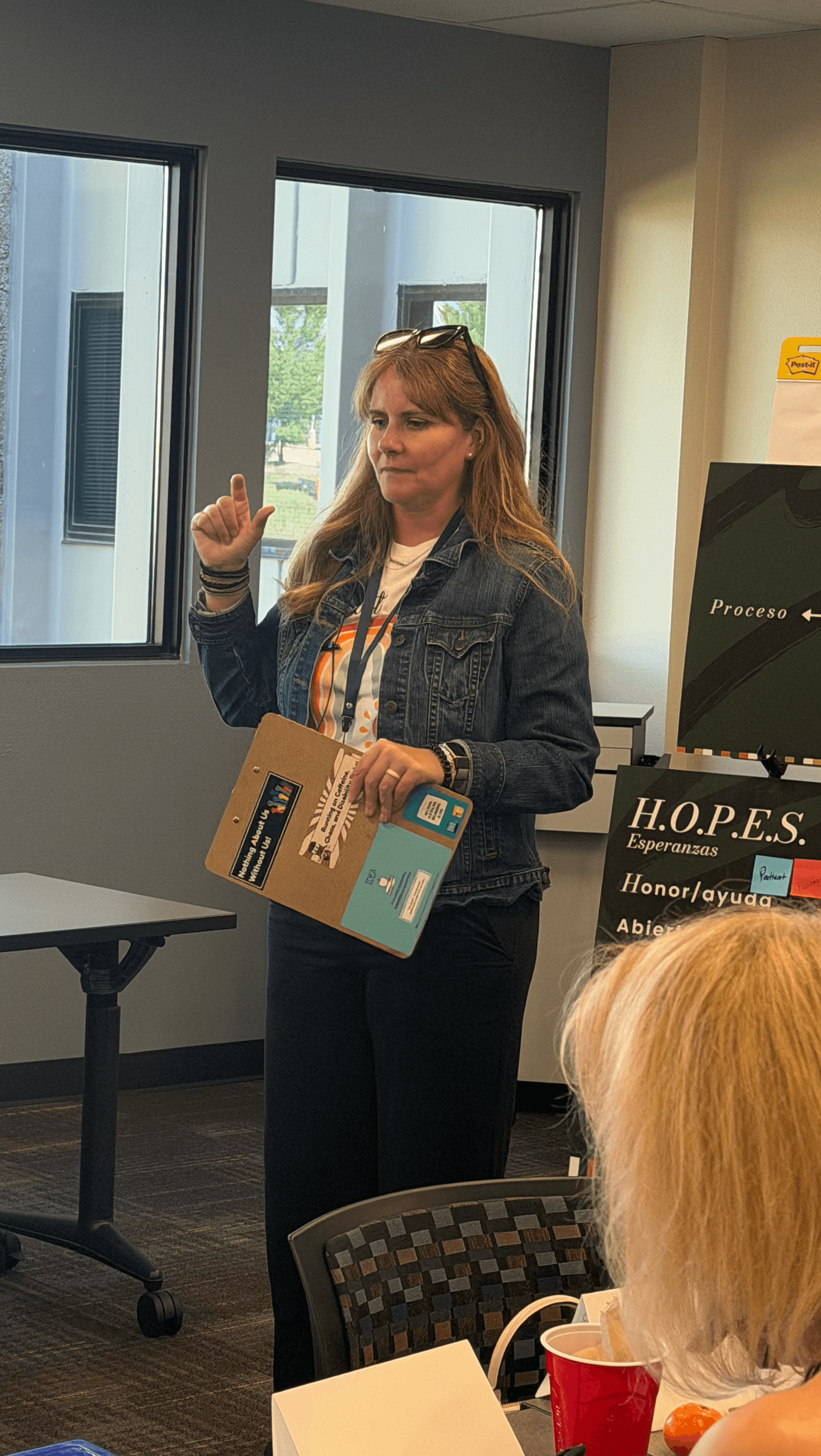 Woman standing in front of windows, holding a box, gesturing with her right hand, in a room with a computer and a person with blonde hair.