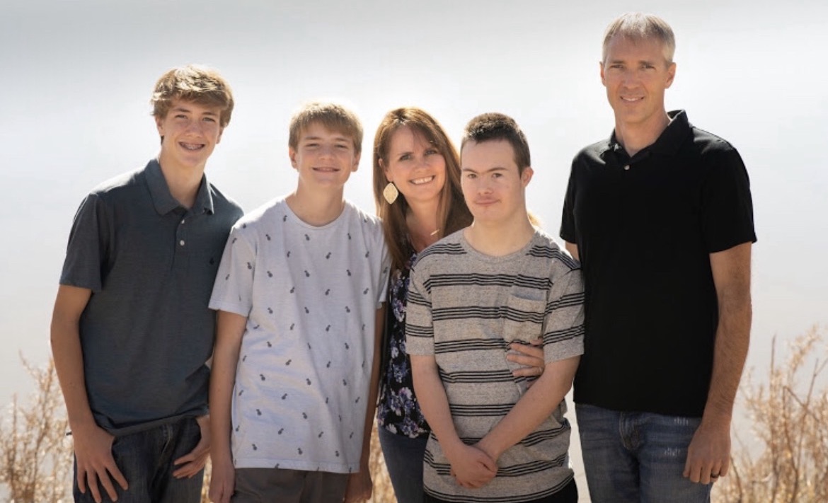 Group of five people standing outdoors, smiling, with a bright sky background.