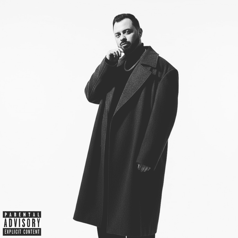 Man with beard in dark coat and turtleneck, standing with hand near face, against white background.
