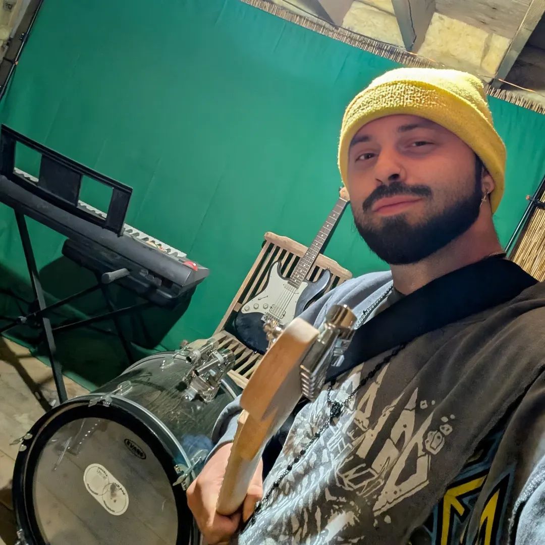 Man wearing yellow beanie holding a guitar, with keyboard and drum set in background, green backdrop behind him.