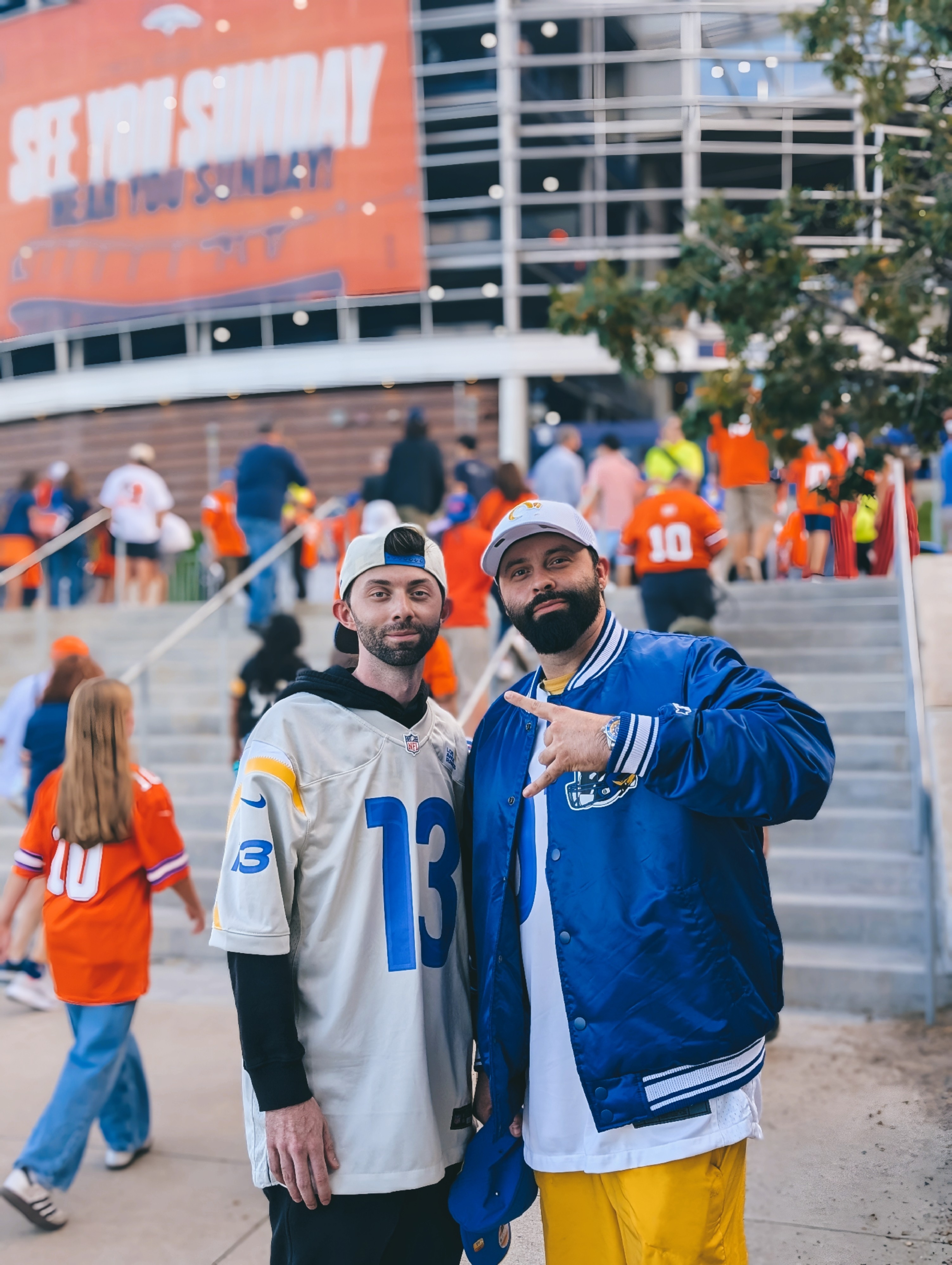 Two men pose outdoors, one in a sports jersey and the other in a blue jacket, with a crowd and stadium in background.