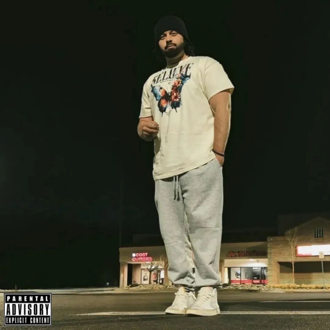 Young man with a beard, wearing a beanie, graphic t-shirt, and sweatpants, standing outdoors at night.
