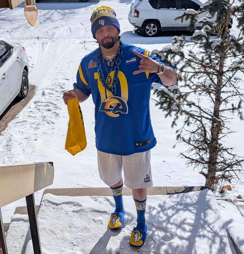 Man in Los Angeles Rams jersey and winter clothing standing outdoors in snow, making a peace sign, holding a yellow item.