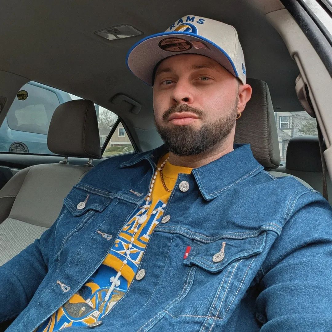 Man with a beard wearing a baseball cap and denim jacket sitting in a car, looking at the camera.