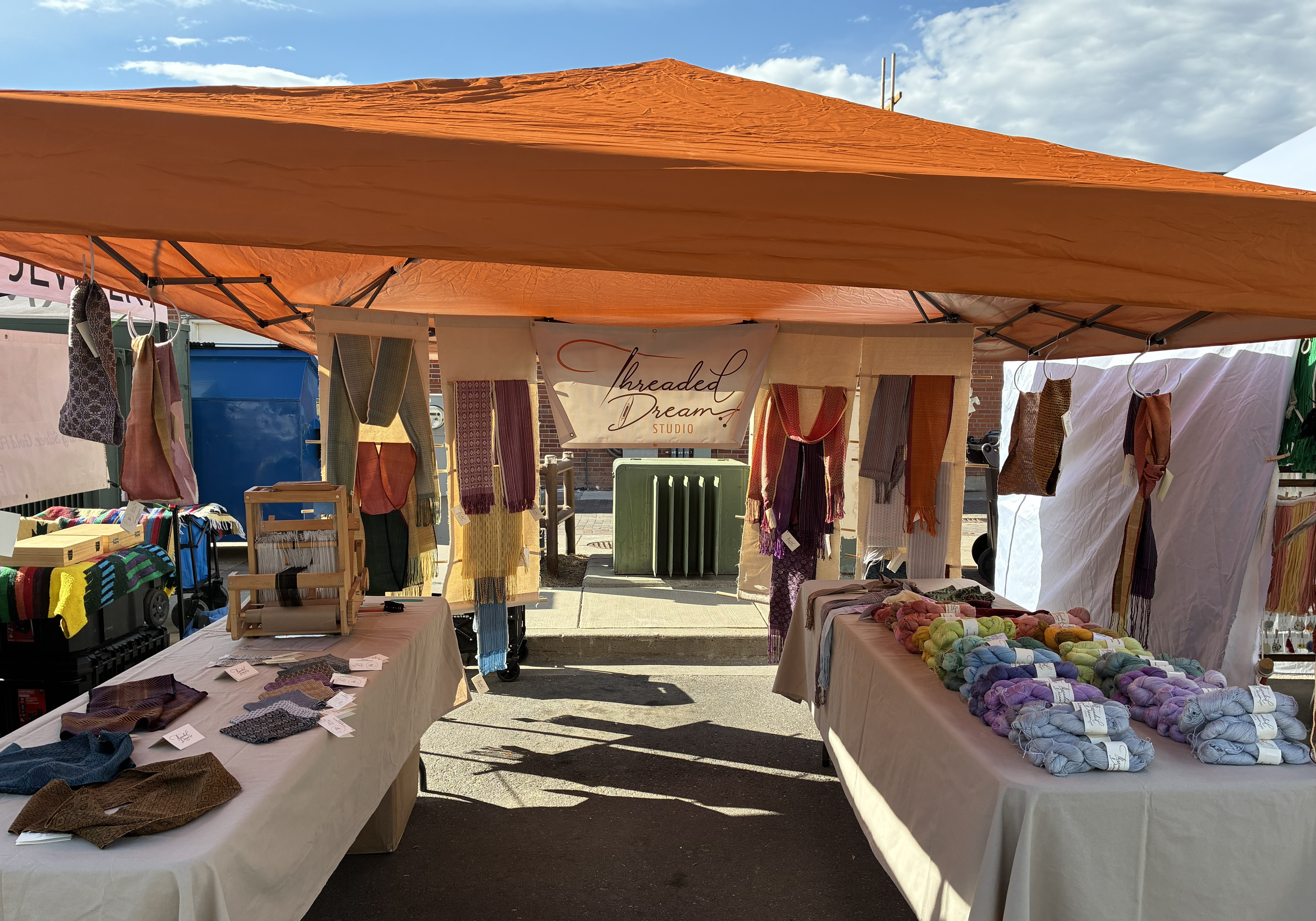 Outdoor market stall with clothing and fabric items displayed on tables and hanging under a canopy, sunny day.