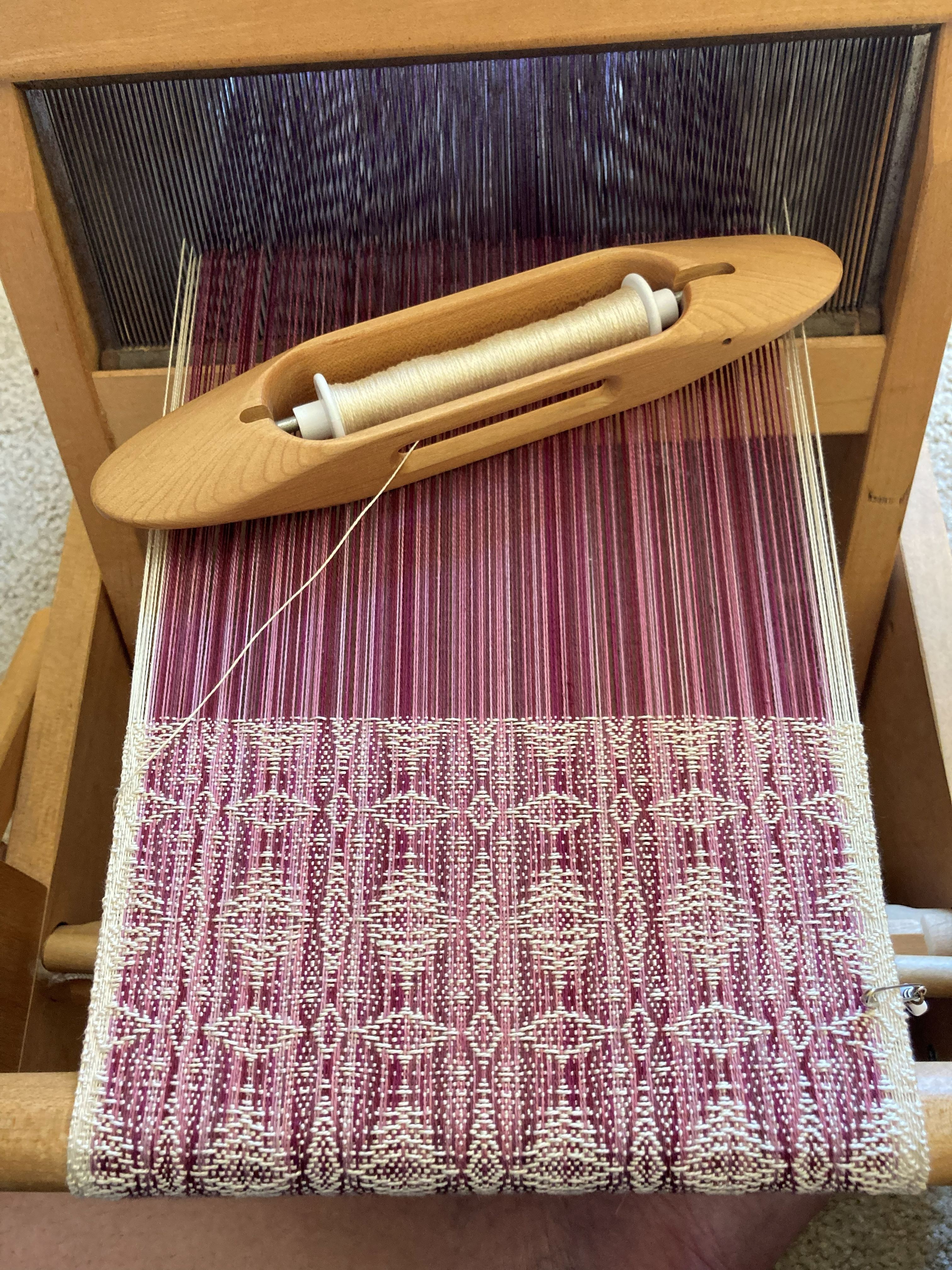 A weaving loom with a partially woven textile in pink, white, and purple colors, and a wooden frame, viewed from above.