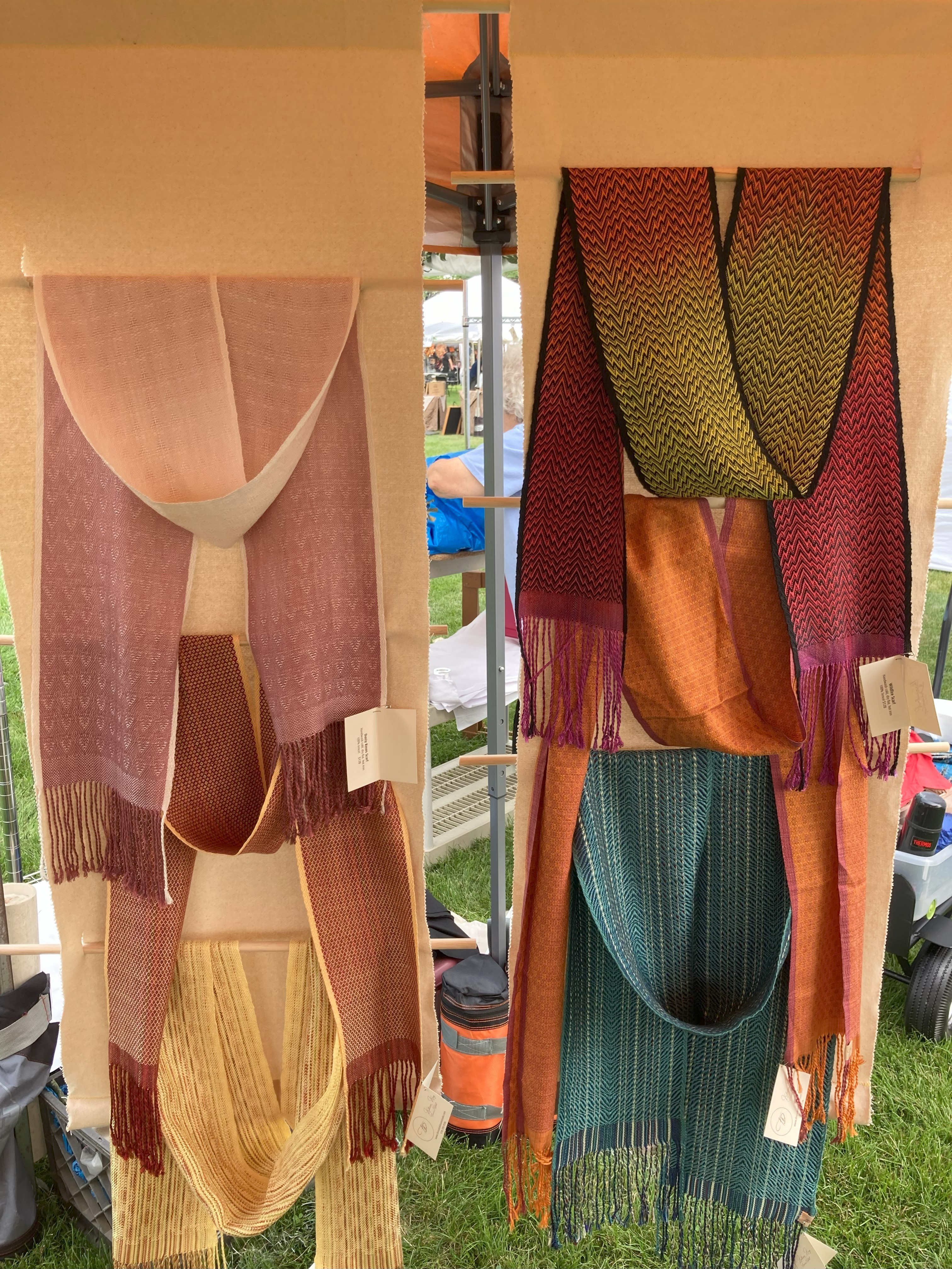 Display of colorful woven scarves hanging vertically, with various patterns and fringes, in an outdoor setting.