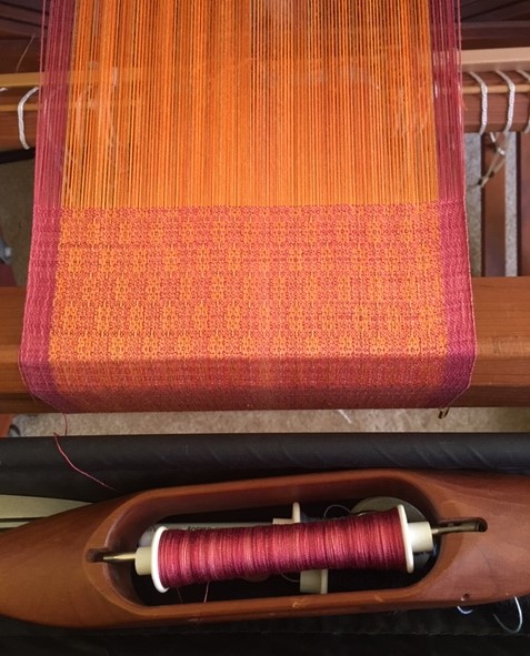 Colorful woven textile on loom with pink and orange threads, and a bobbin of pink thread below. Word count: 20
