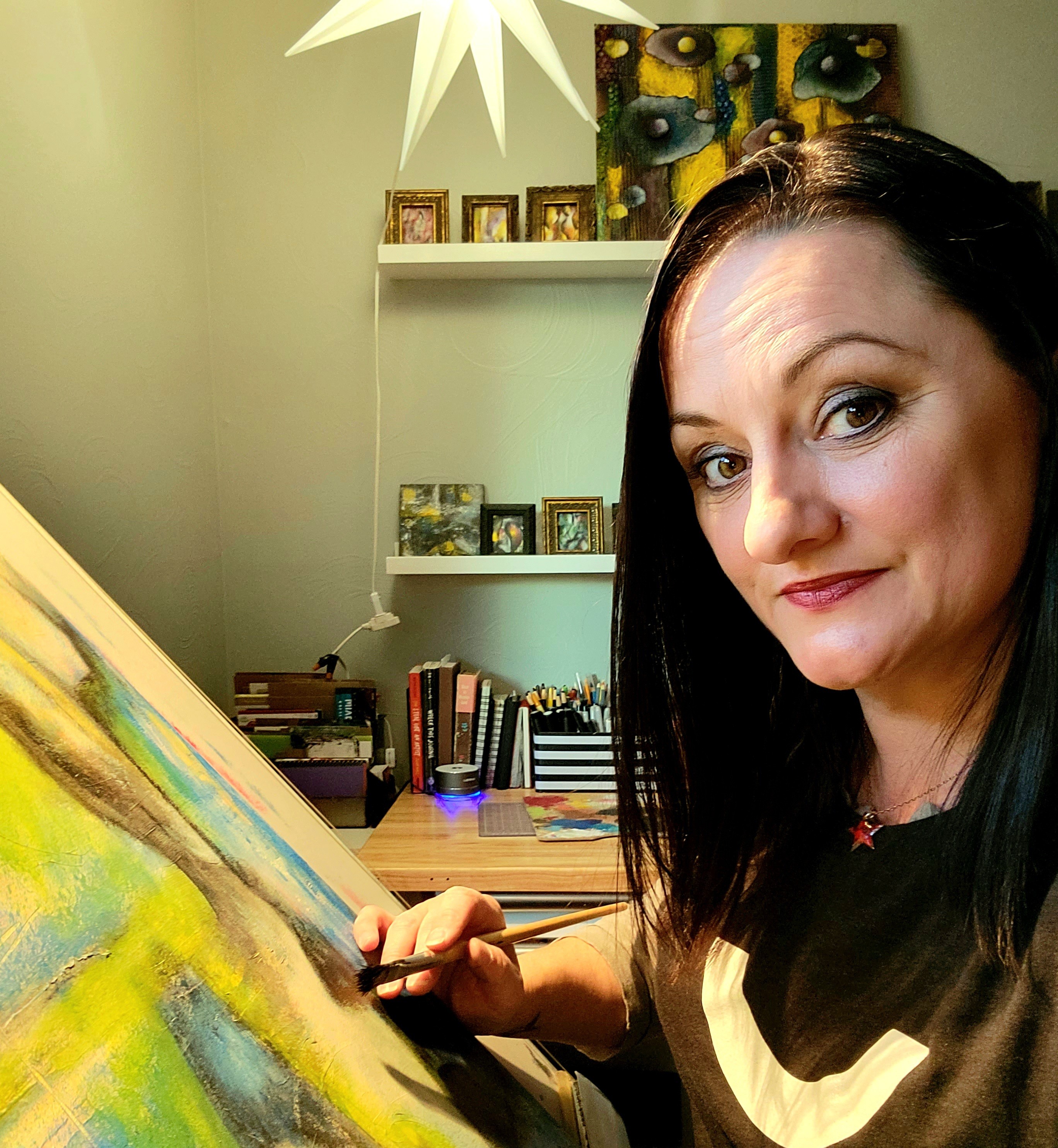 Woman with black hair painting on canvas in a room with shelves and artwork behind her.