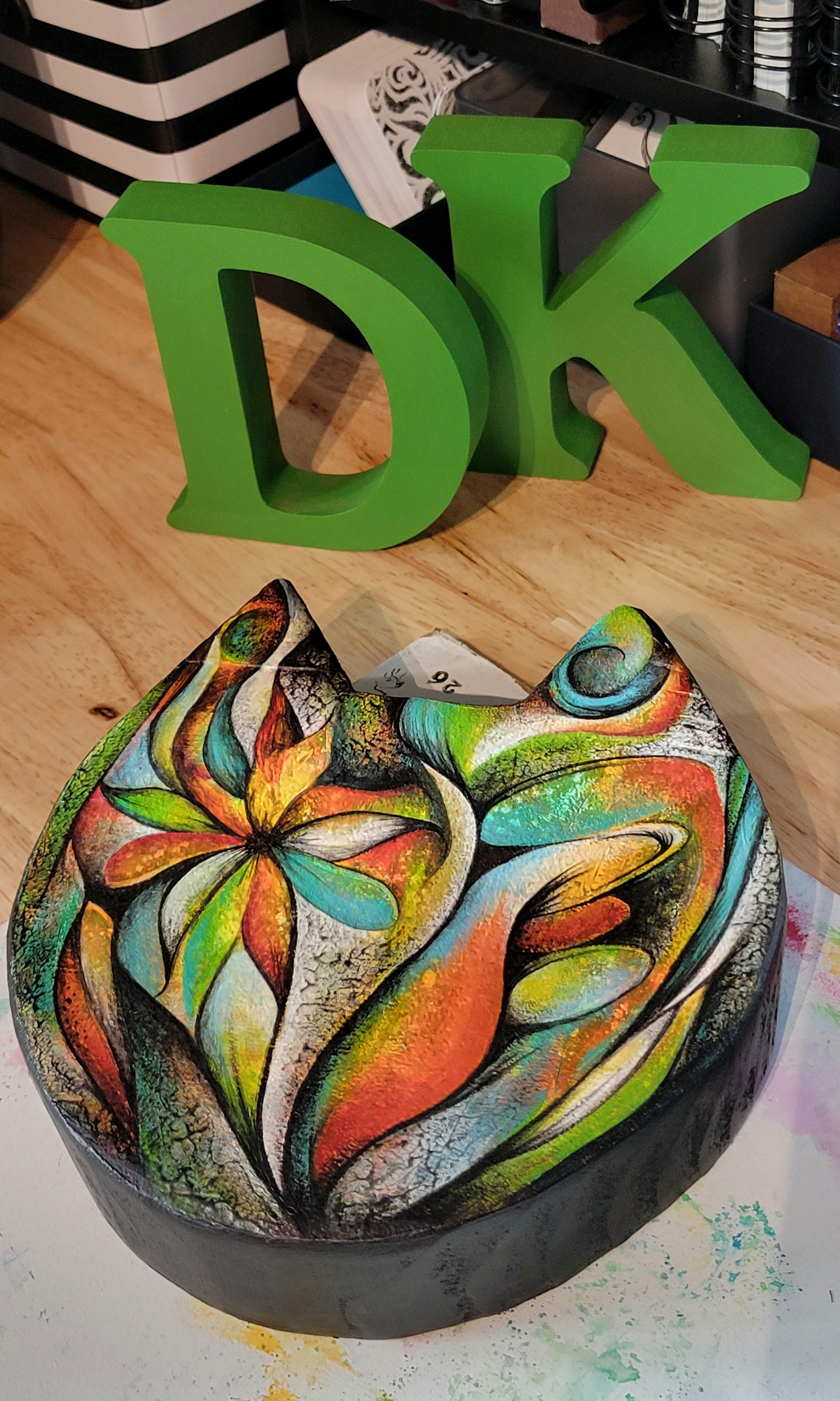 Colorful, abstract painted heart-shaped object on a table with green letters 'D' and 'K' behind it.