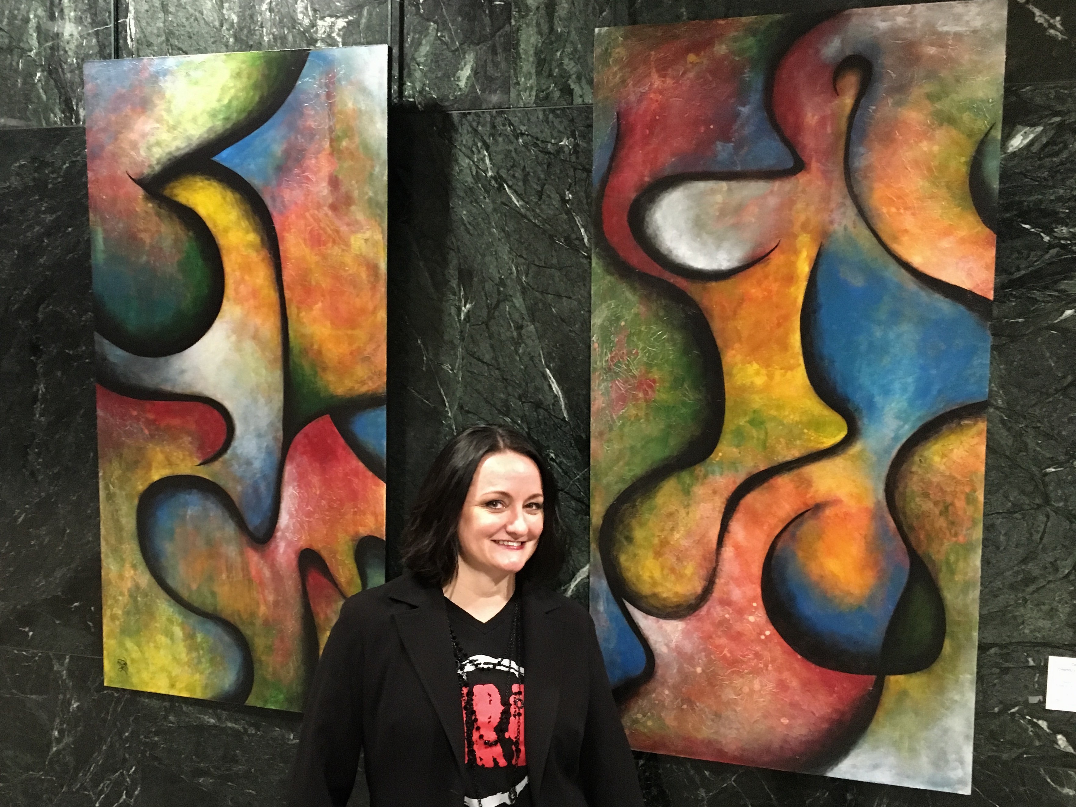 Woman with dark hair smiling in front of colorful abstract paintings on a dark wall.