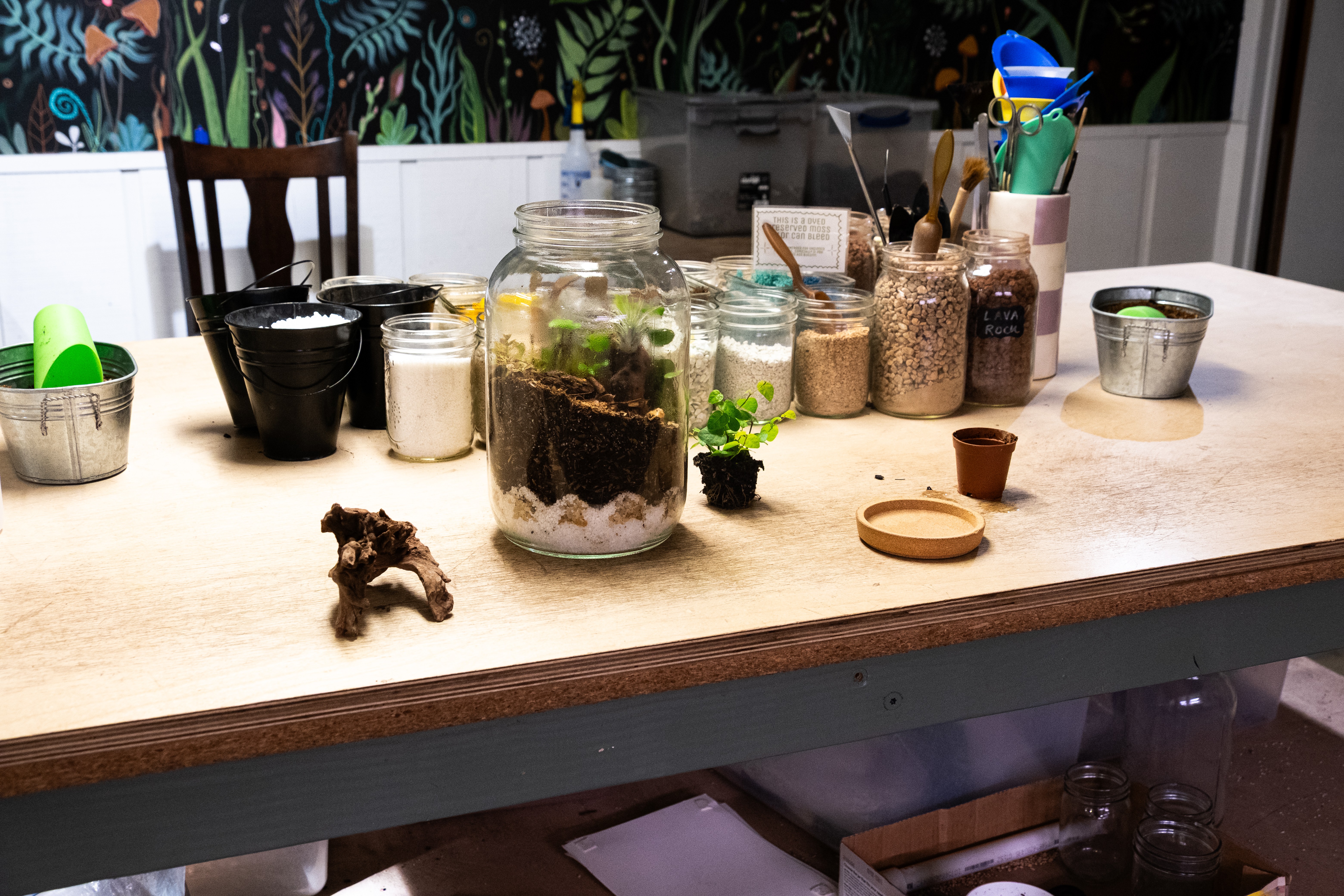 Table with jars, containers, and gardening supplies, with chairs and a colorful patterned wall in the background.
