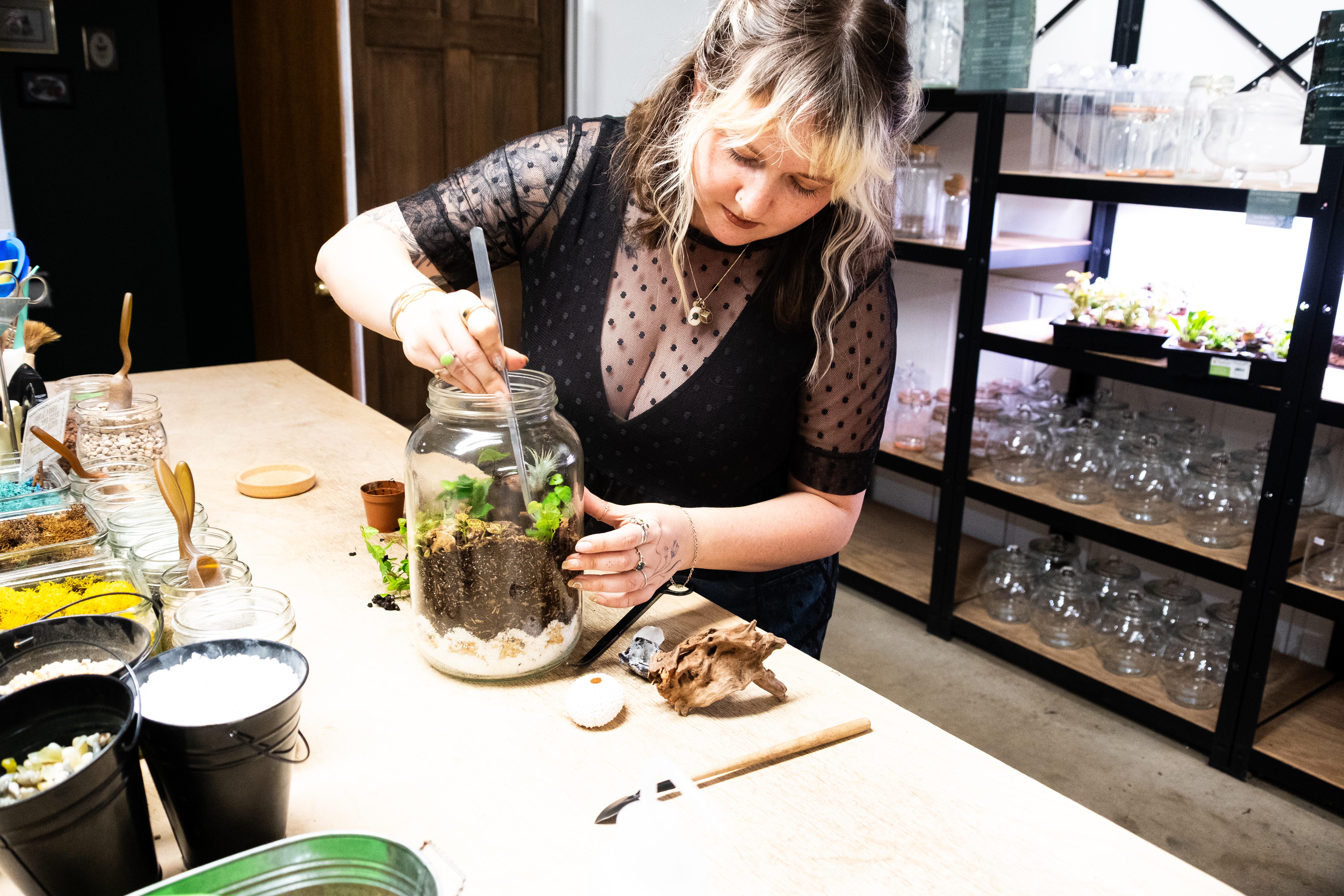 Woman tending to a small plant in a glass container on a table, with gardening tools and supplies nearby.