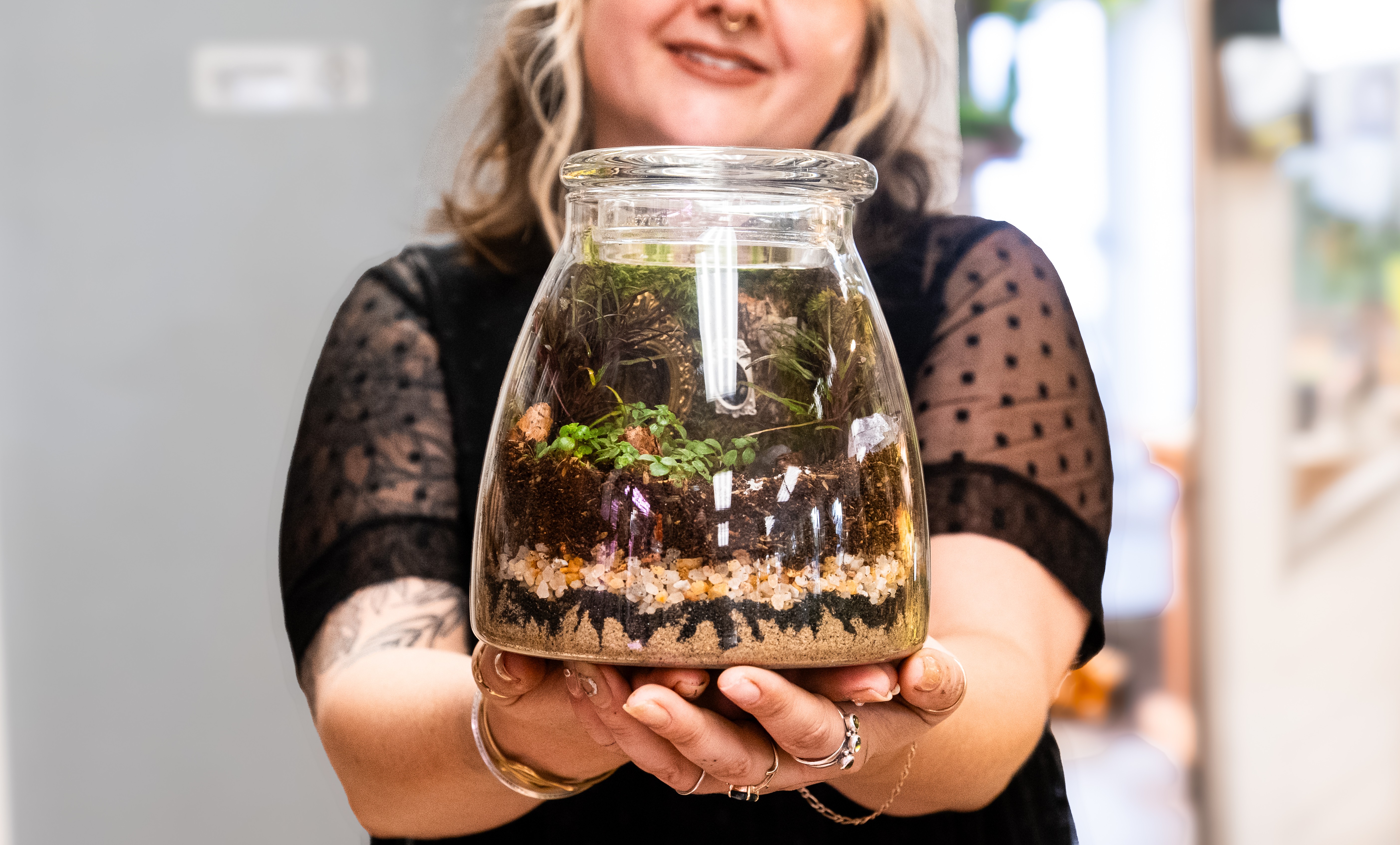 Person holding a glass jar with plants and soil inside, smiling, wearing a black top with sheer sleeves.