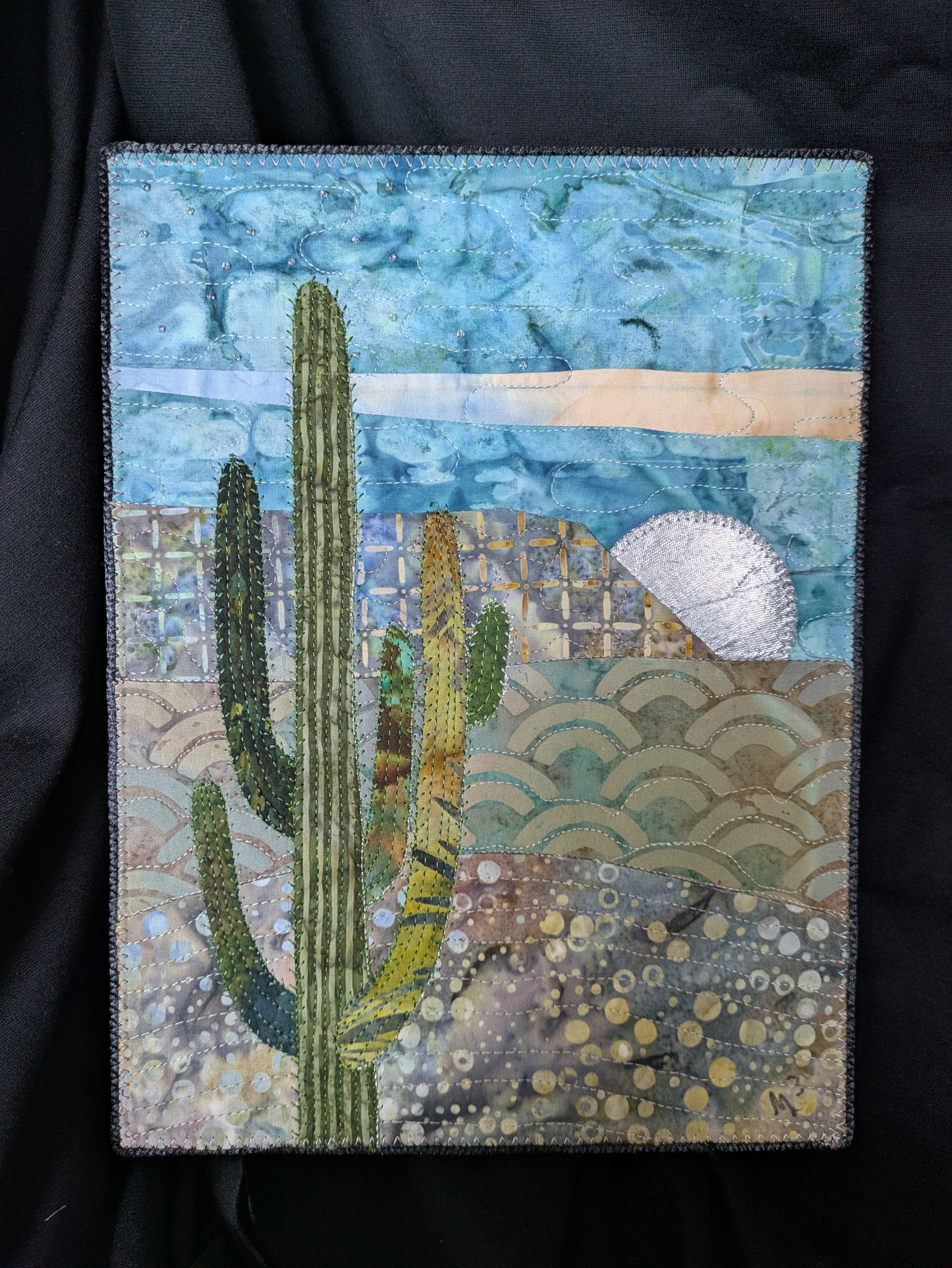 Fabric patch depicting a desert landscape with cacti, a setting sun, and stylized clouds and waves, in vibrant colors.