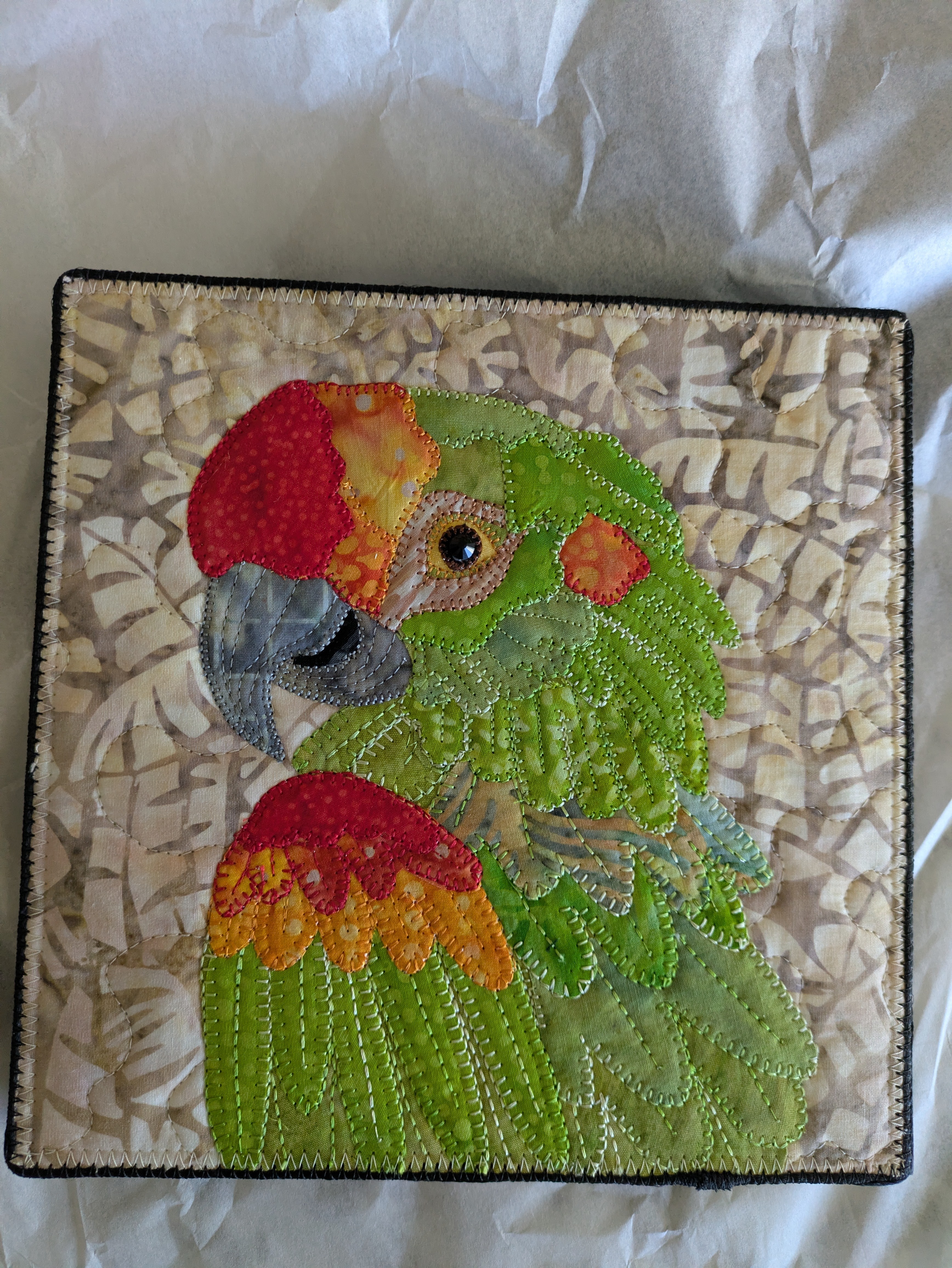 Colorful embroidered parrot with red, green, yellow, and orange feathers on a textured background.