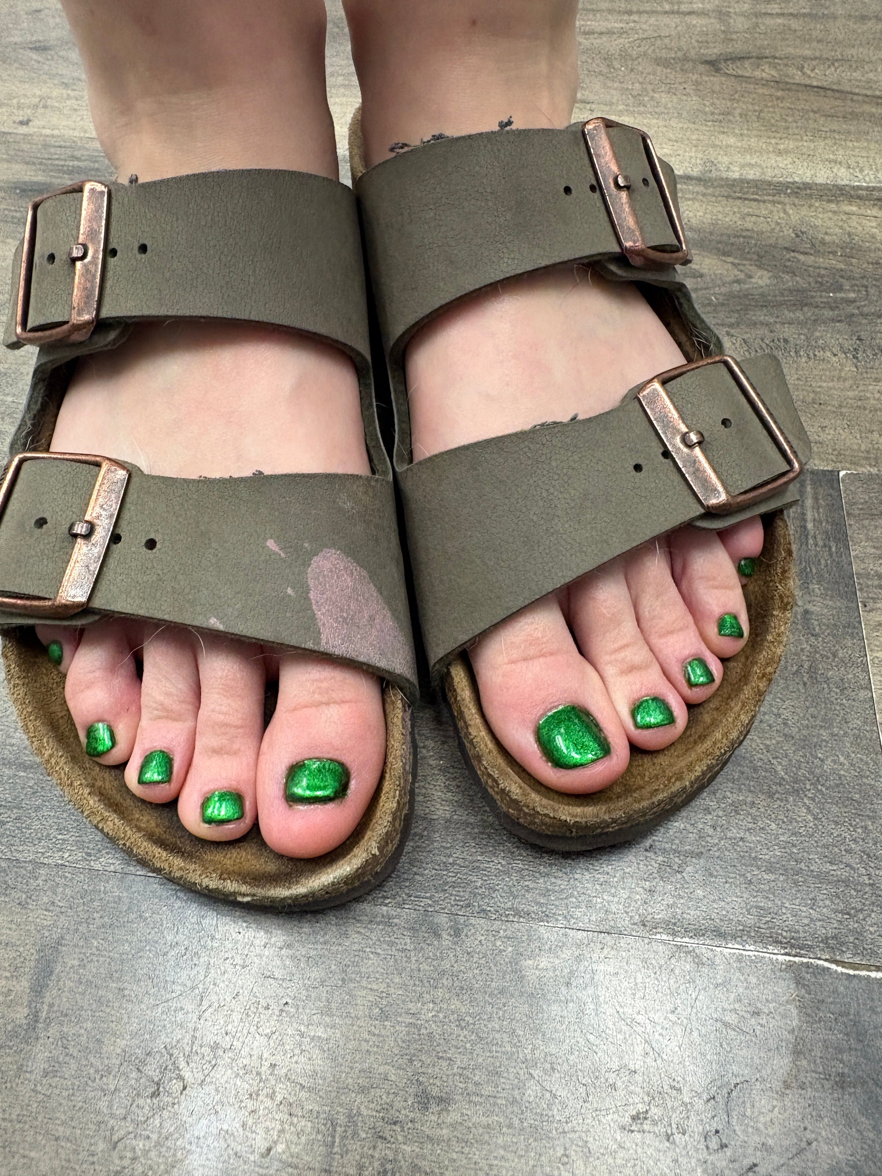 Person wearing green toenail polish and sandals with thick straps and buckles, standing on a gray tiled floor.
