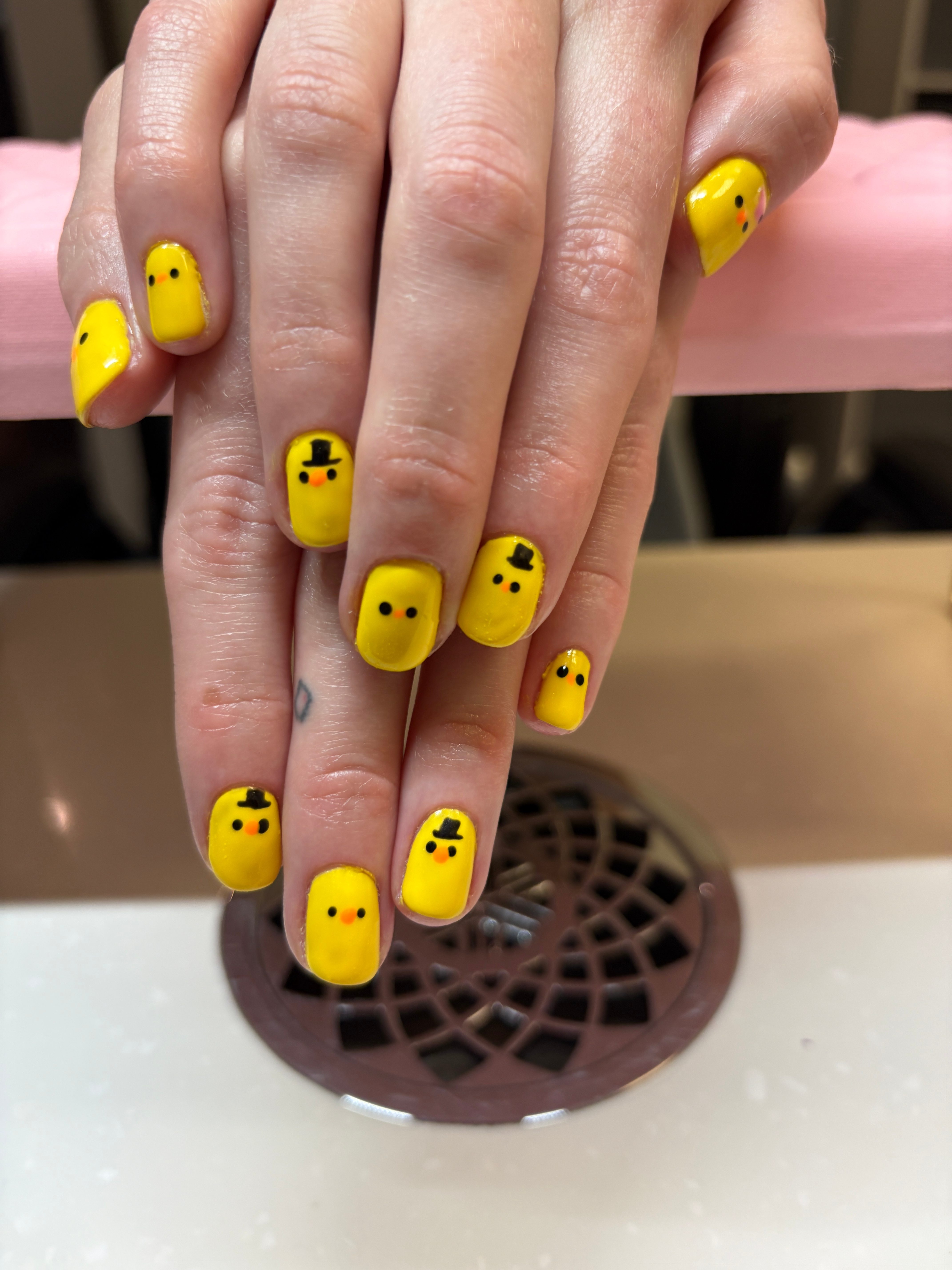 Hands with yellow nail polish and black smiley face designs near a white surface and a brown vent, fingers overlapping.