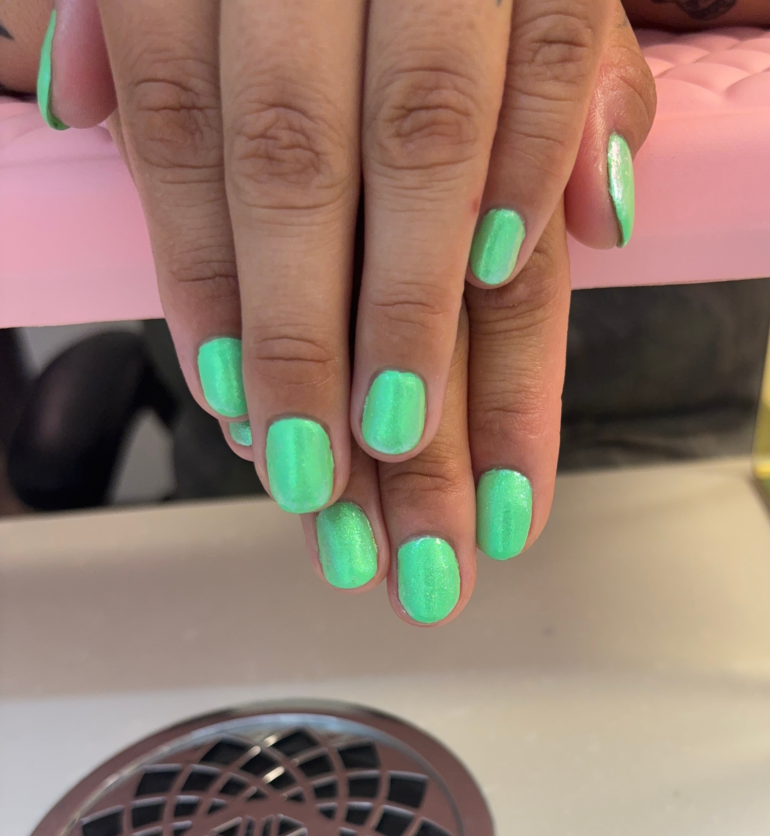 Hand with nails painted in bright green, resting on a pink surface, with a white surface and a circular vent below.
