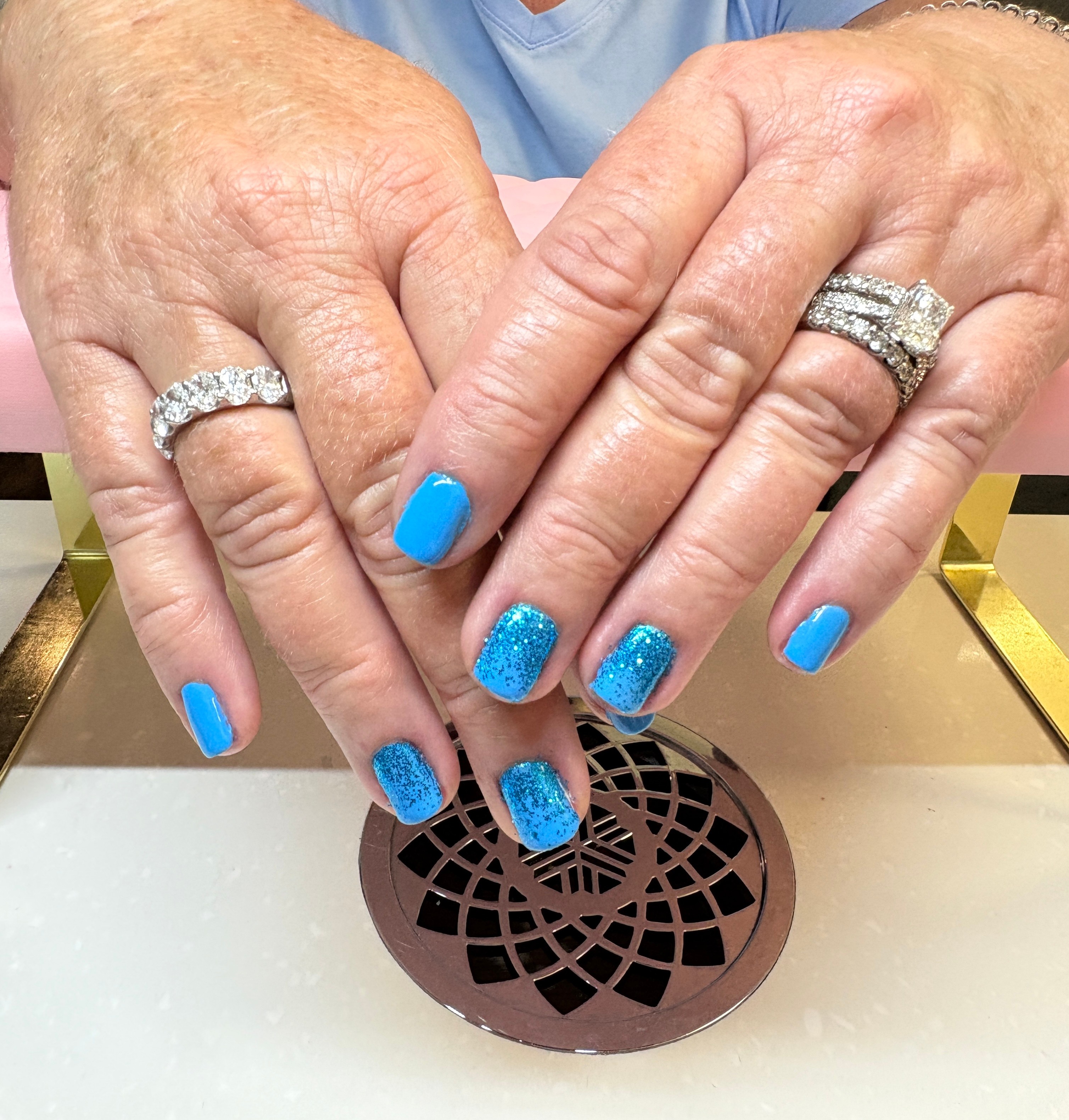 Two hands with rings and blue glittery and solid blue nail polish resting on a sink drain.
