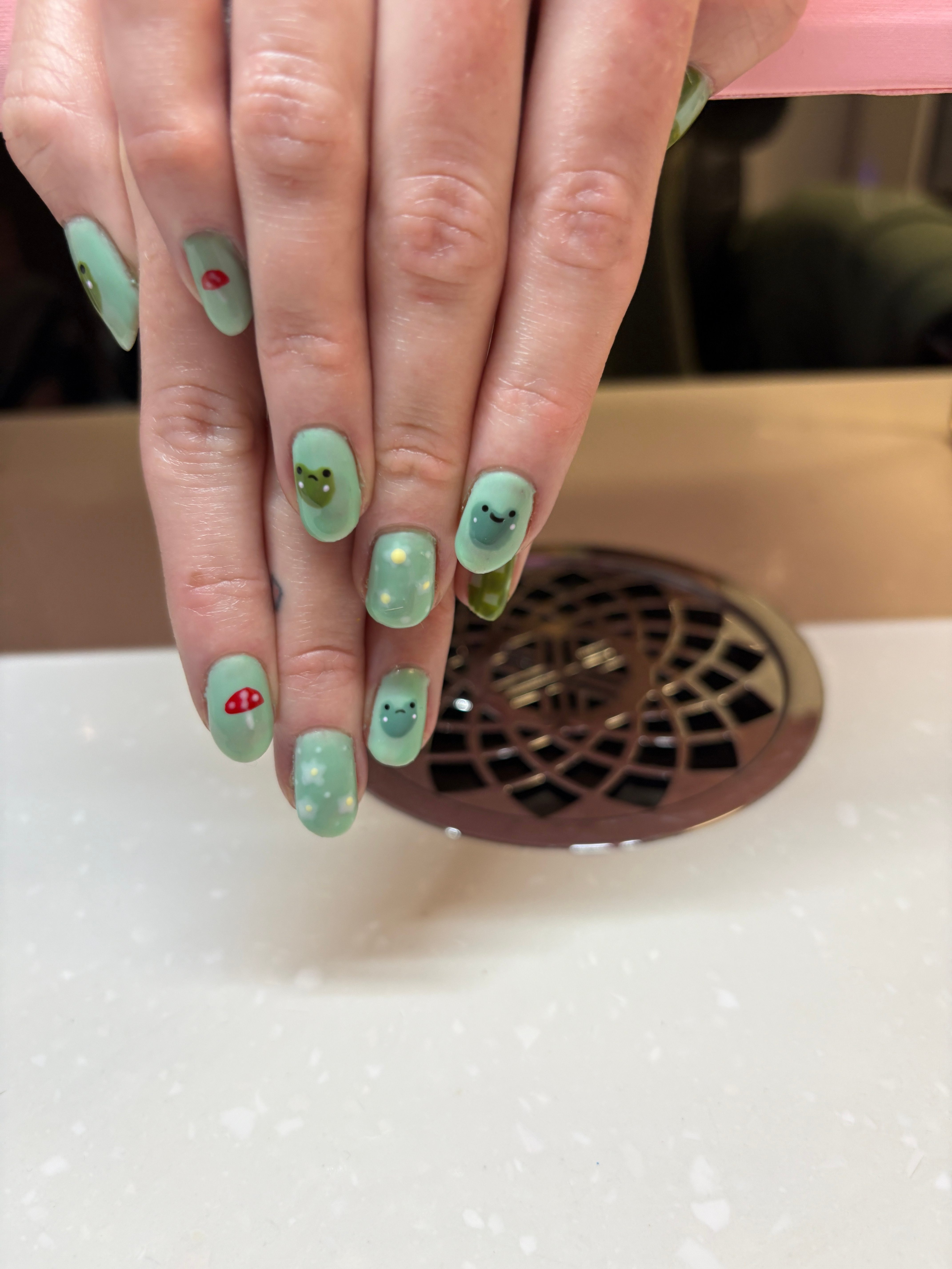 Hand with mint green nails and decorative designs, near a circular vent on a white surface.