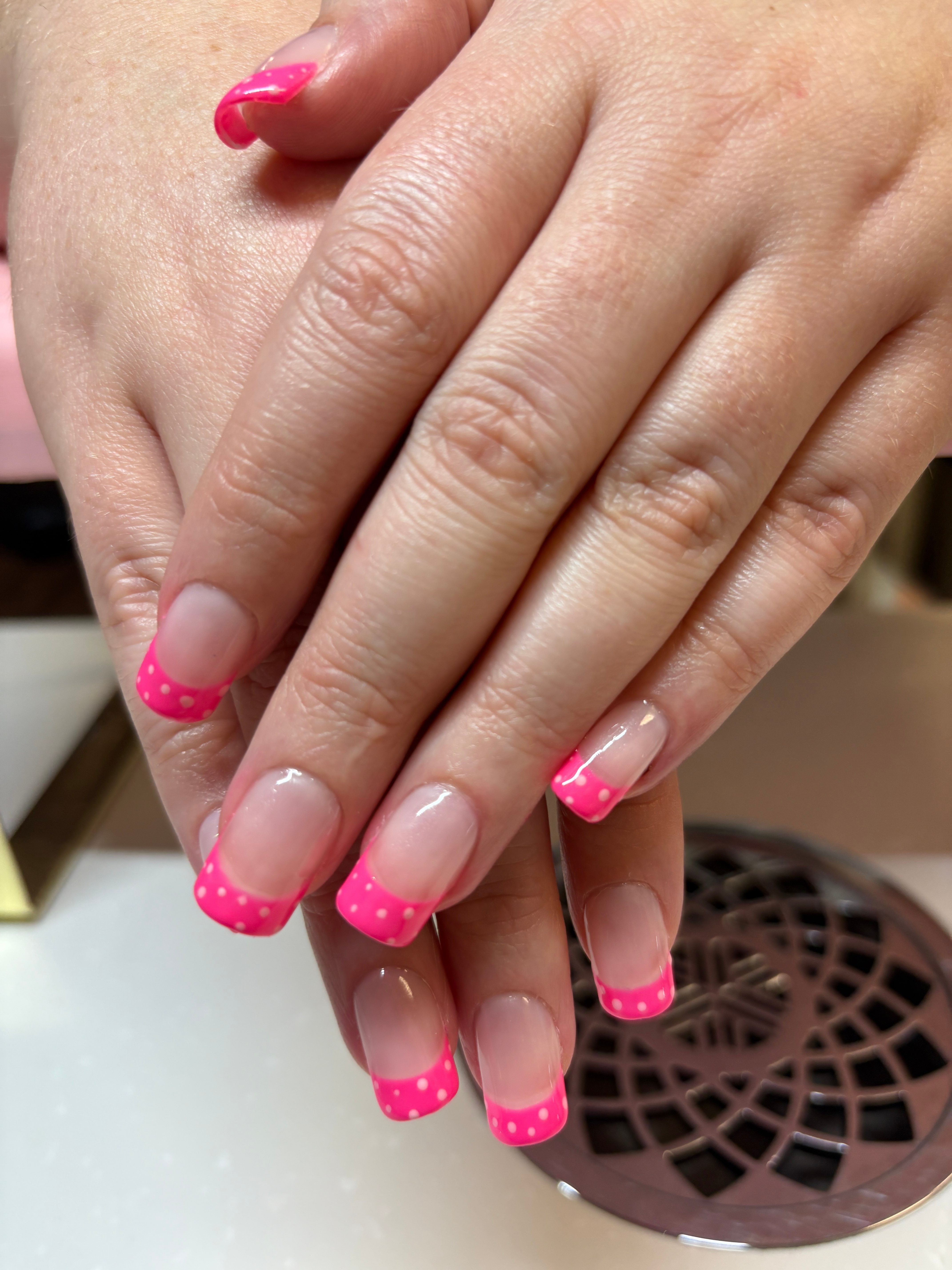 Hands with pink and white polka dot nail polish resting on a surface, with a fan in the background.