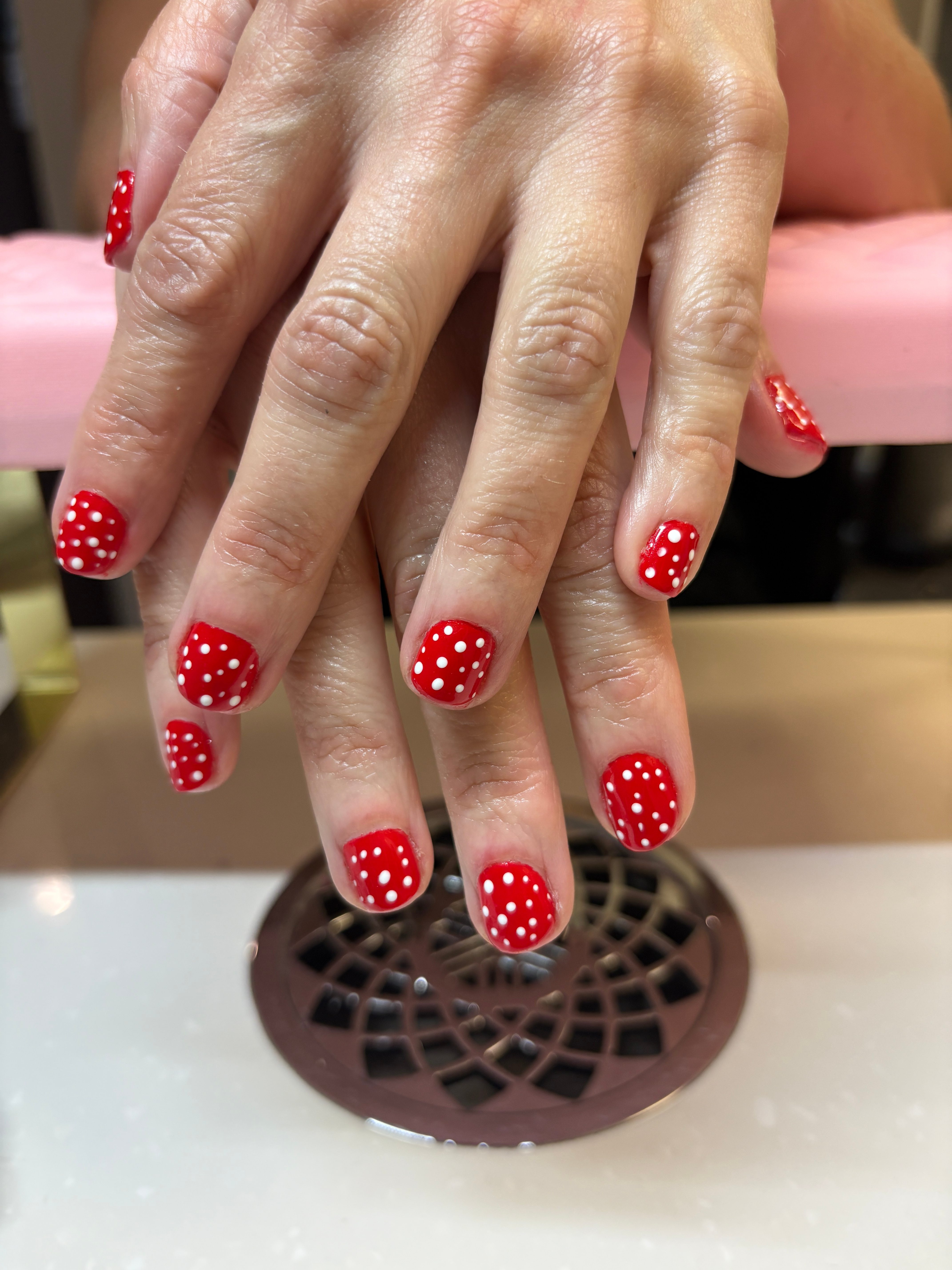 Hands with red polka dot nail polish near a vent, holding a pink drink, background blurred.