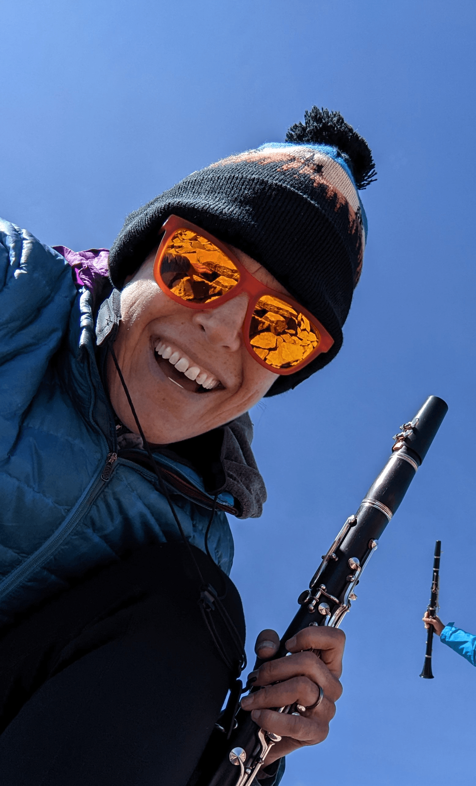 Person smiling, wearing sunglasses and a beanie, holding a clarinet outdoors against a clear blue sky.