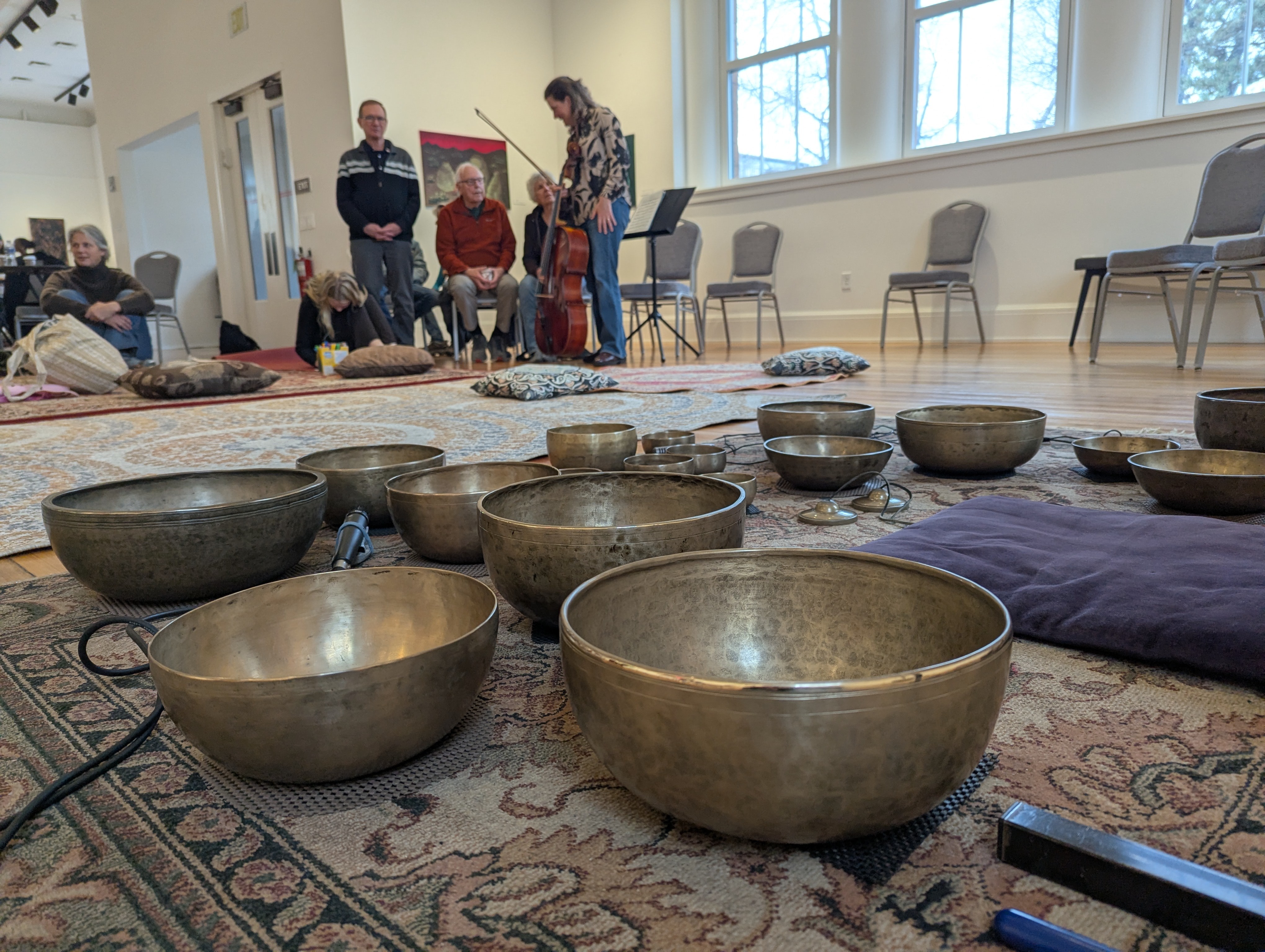 Multiple singing bowls on a patterned rug with people sitting and standing in a room with large windows.