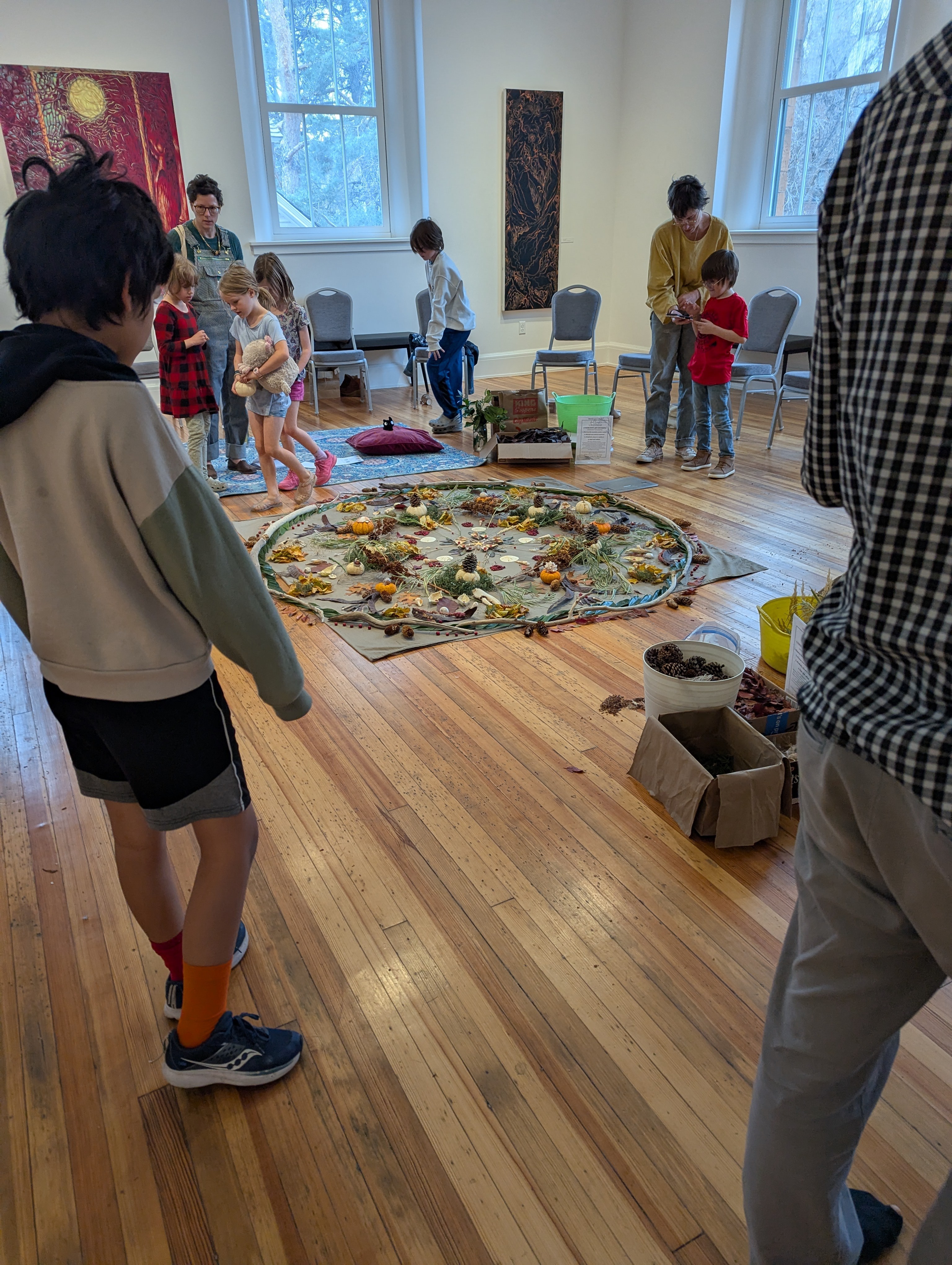 Children and adults gather around a colorful rug in a bright room with large windows, chairs, and toys.
