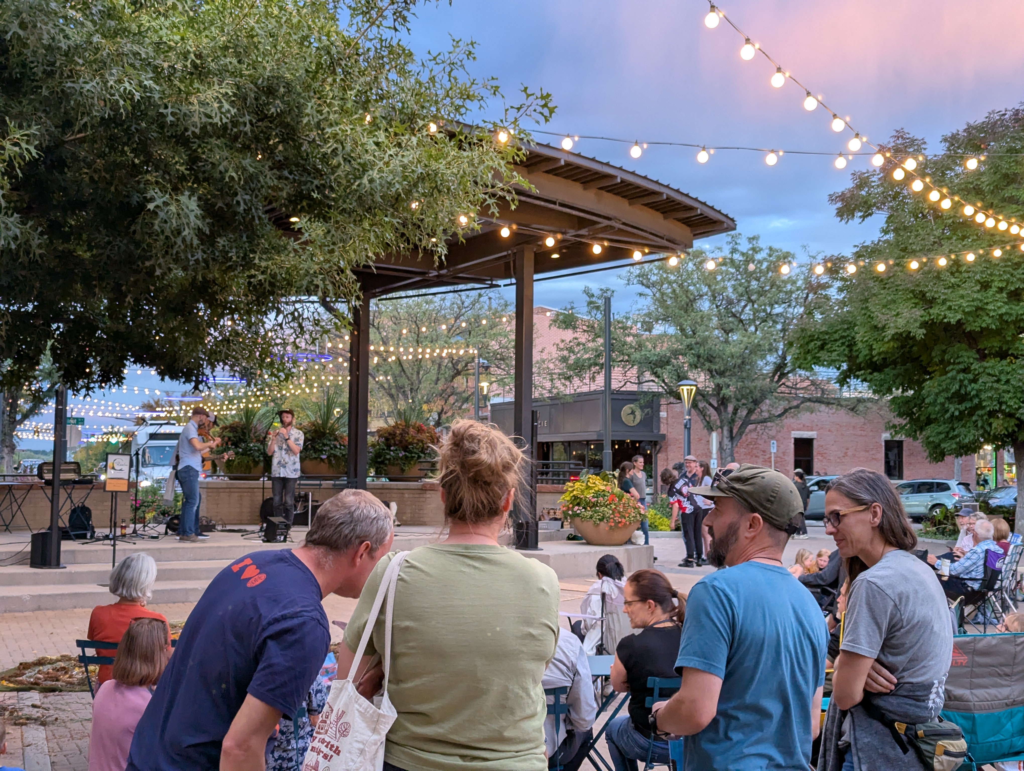 People gather outdoors under string lights, watching a performance on a small stage with trees and buildings in the background.