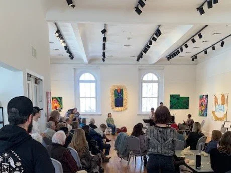 People seated in an art gallery watching a presentation, with paintings on white walls and large windows.