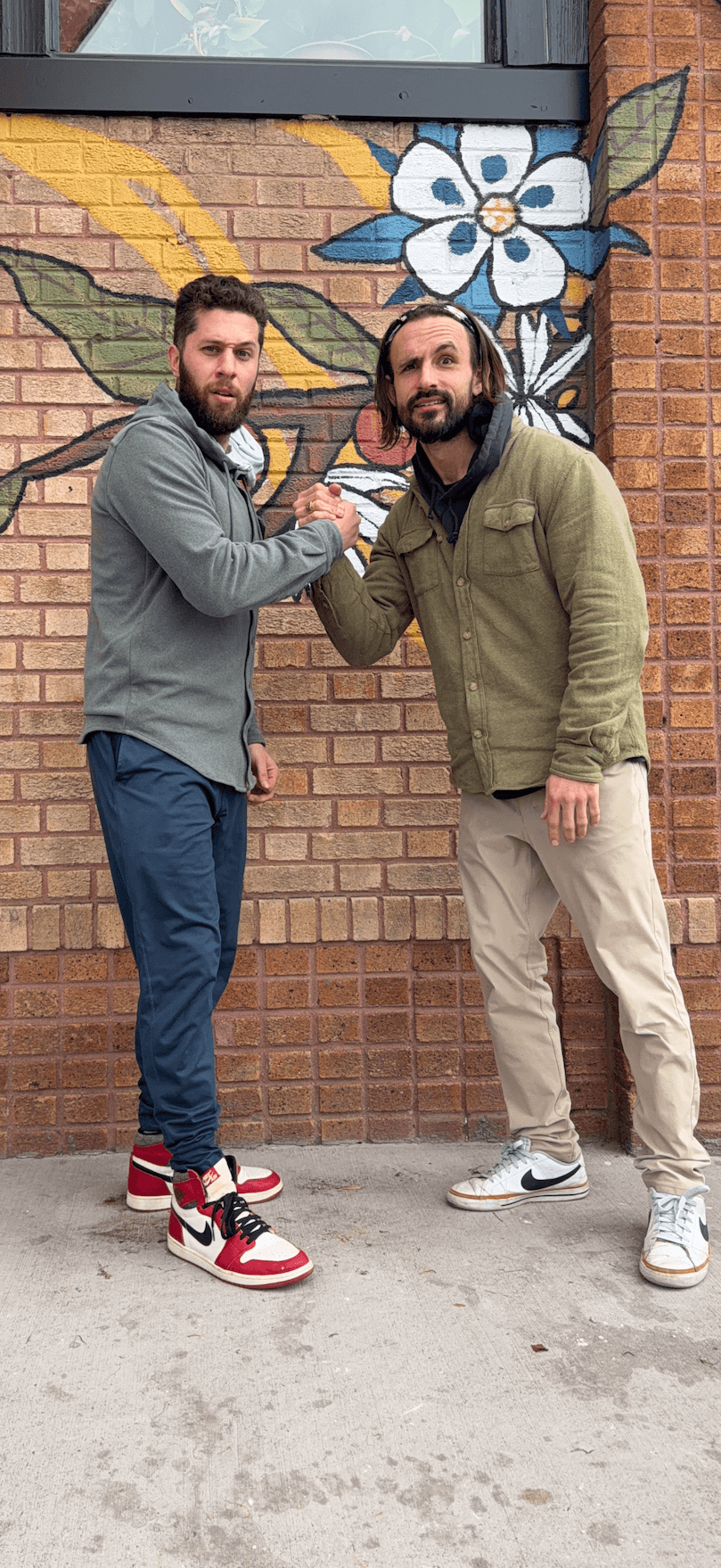 Two men shake hands in front of a brick wall with a colorful flower mural, one wearing a gray jacket and red sneakers.