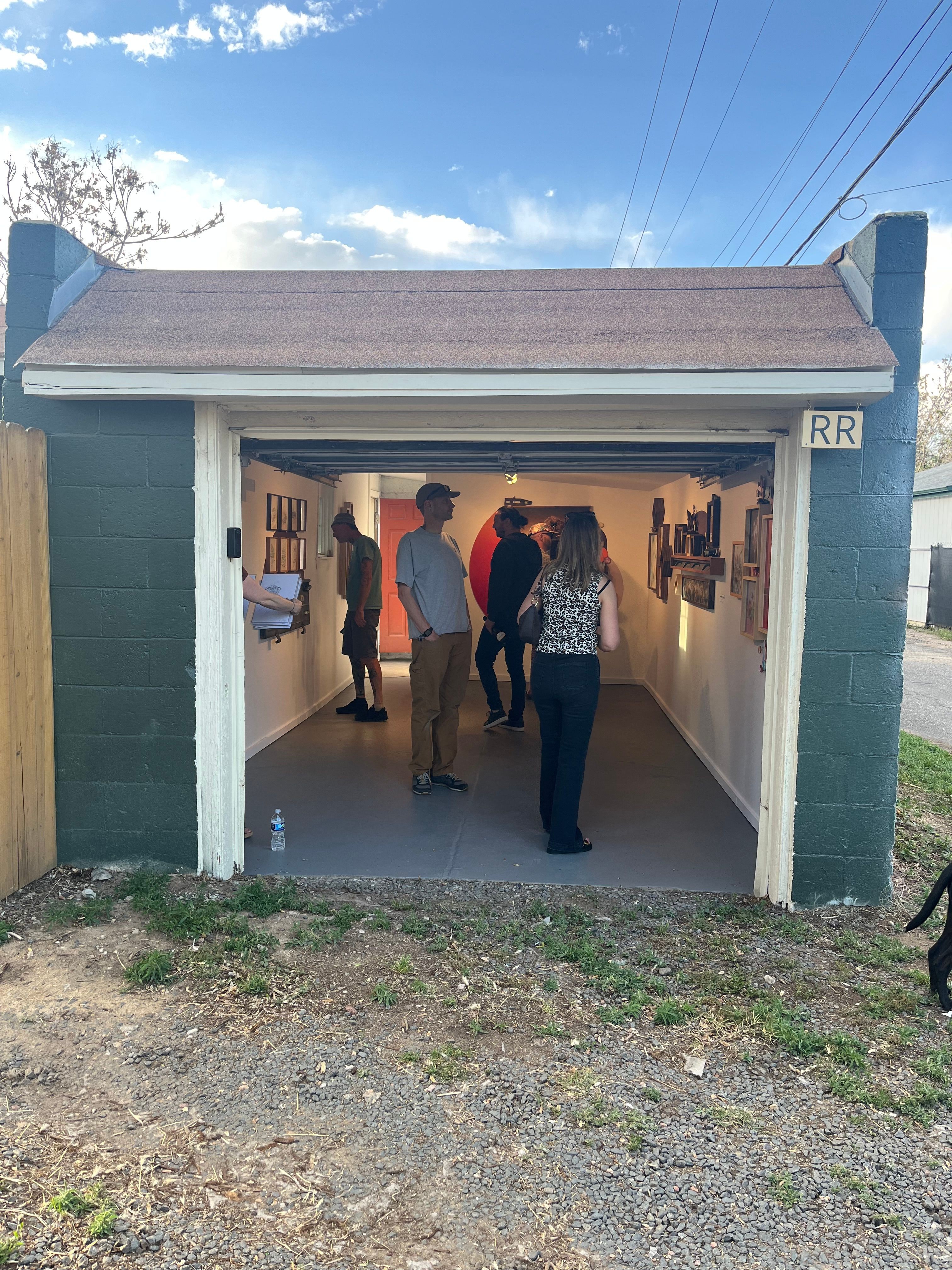 People inside a small building with an open door, some standing and talking, with a person sitting at a table inside.