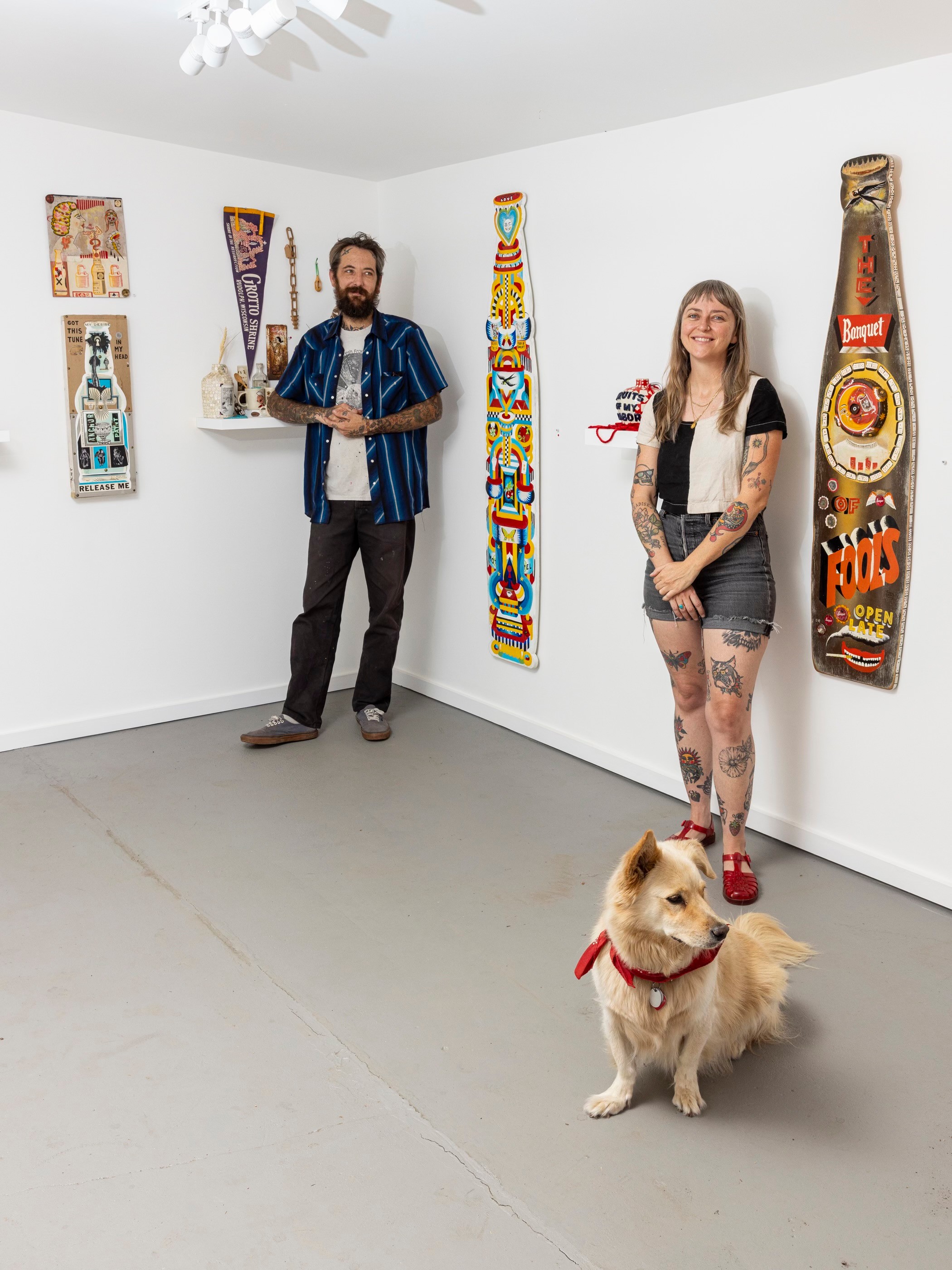 Two people and a dog in an art gallery with colorful vertical artworks on white walls.
