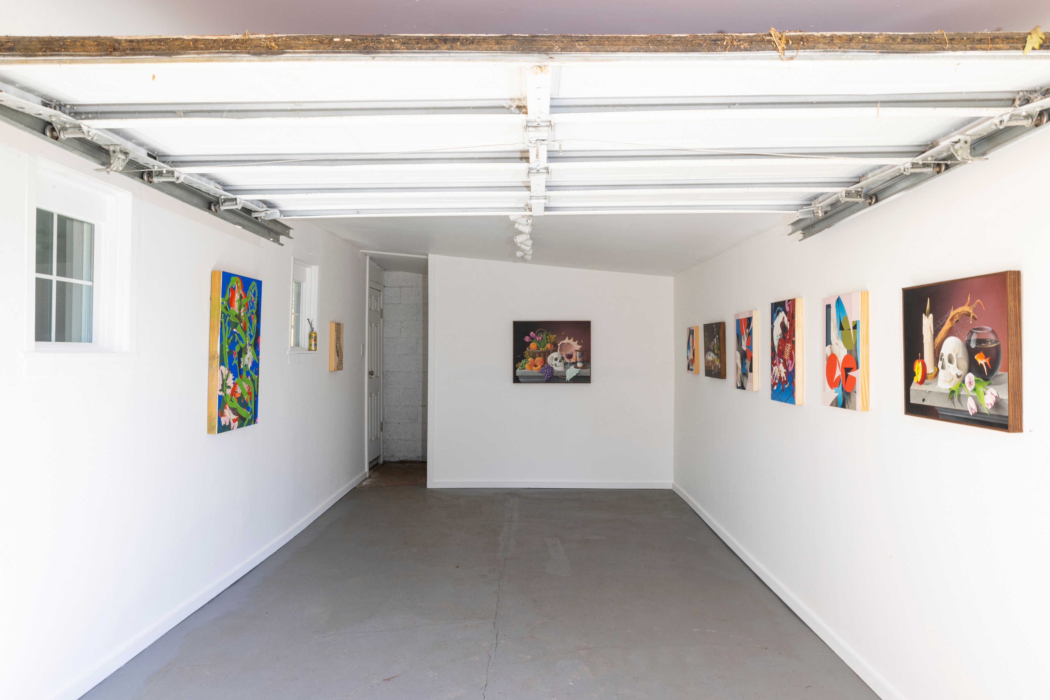 Gallery with white walls and colorful paintings displayed, gray floor, and ceiling with exposed beams.