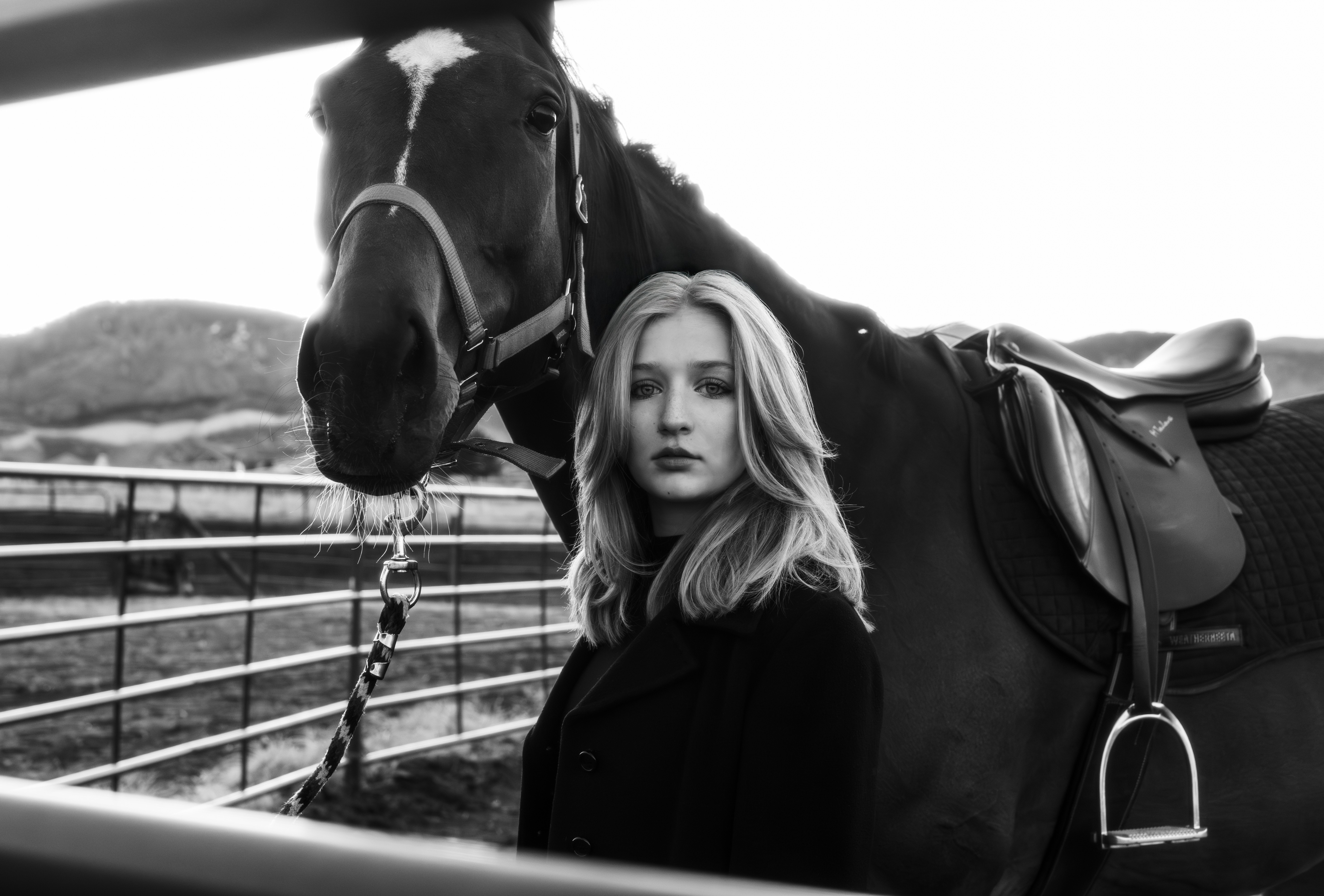 Young woman with shoulder-length hair standing next to a horse, both behind a fence, outdoors in black and white.