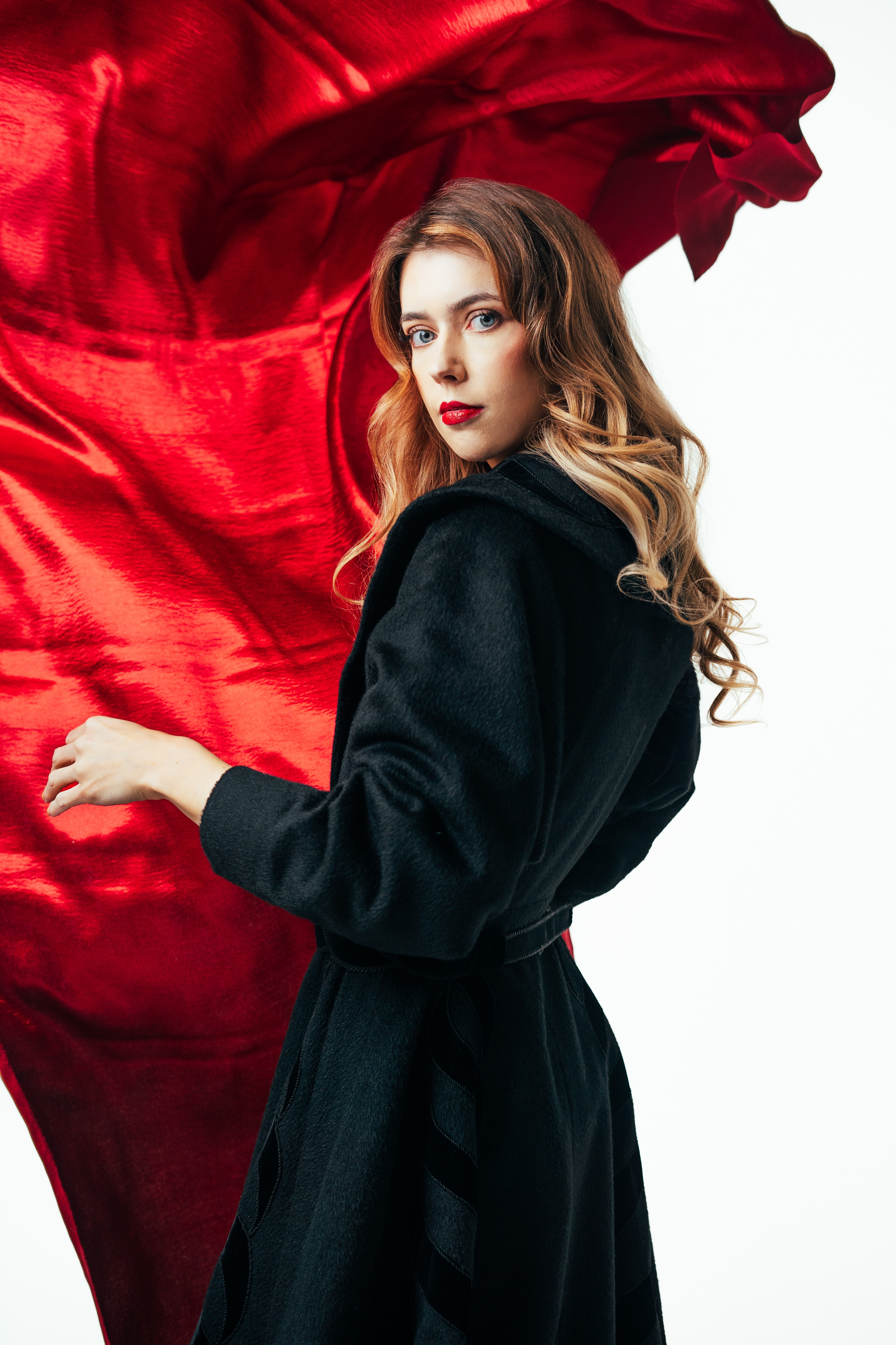 Young woman with long wavy hair wearing a black coat, standing against a white background with red fabric behind her.