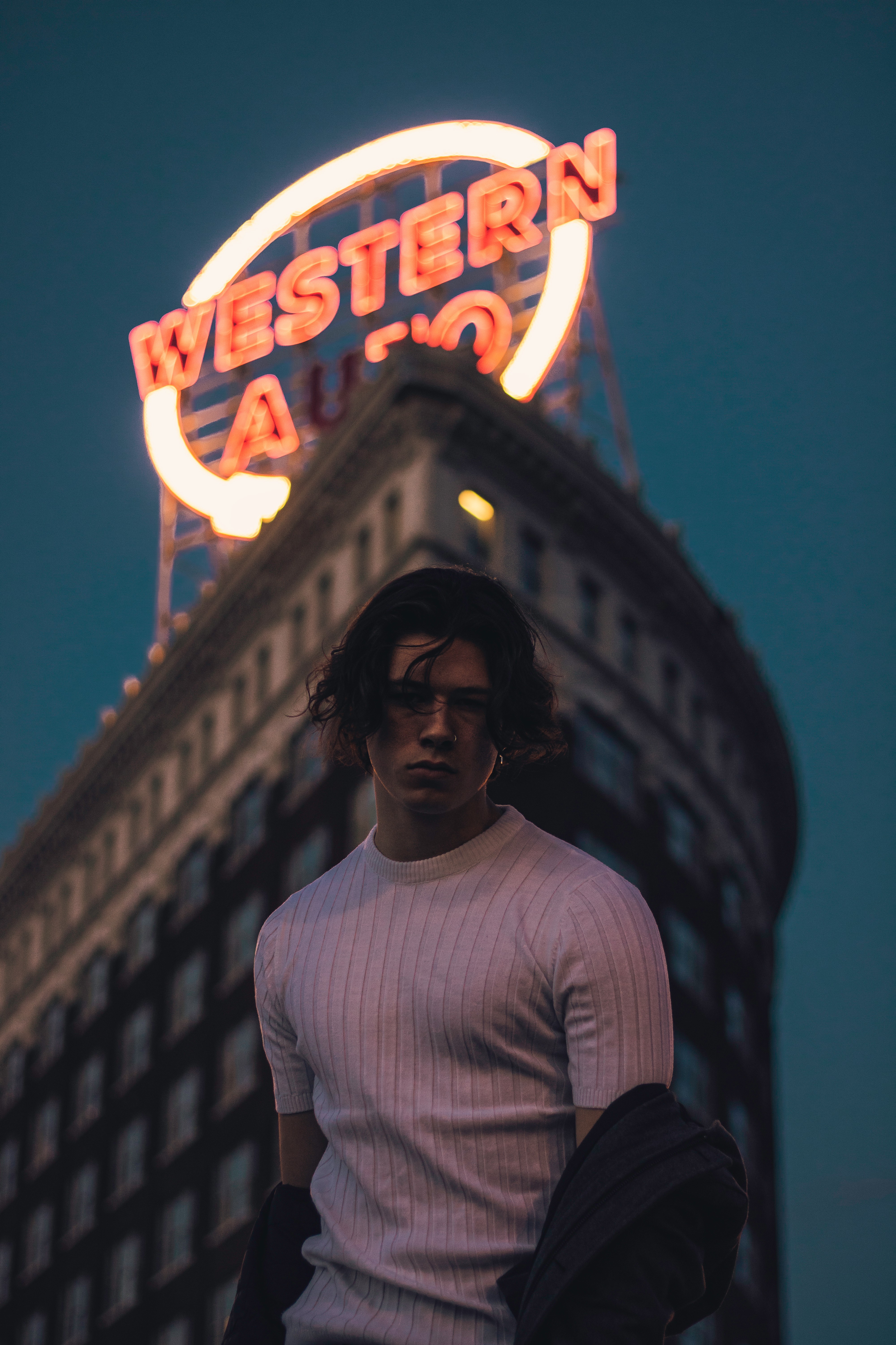 Person with dark hair and glasses wearing a white sweater stands in front of a building with a neon sign reading 'Western' at night.