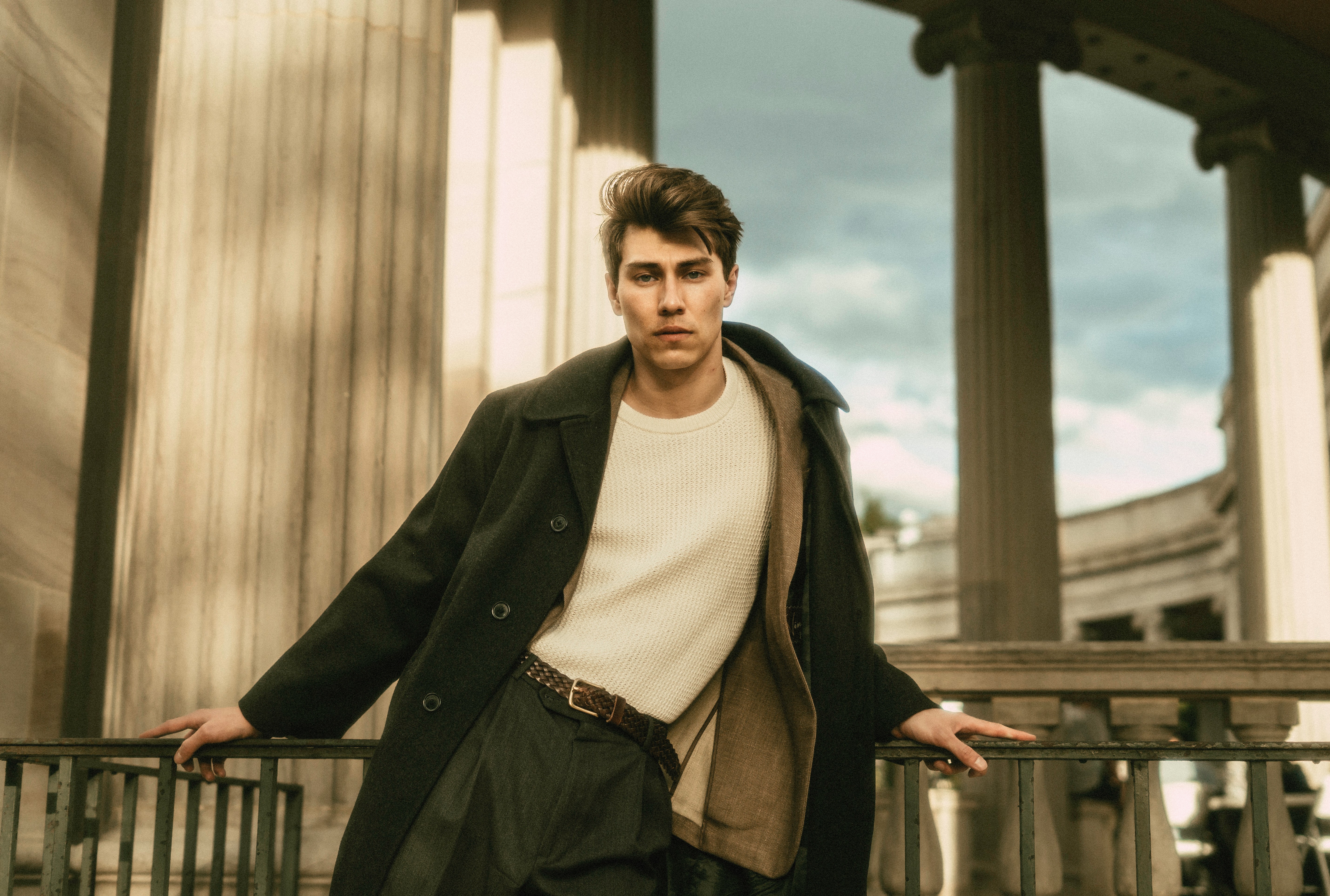 Young man standing outdoors with columns and a railing, wearing a coat and looking at the camera.